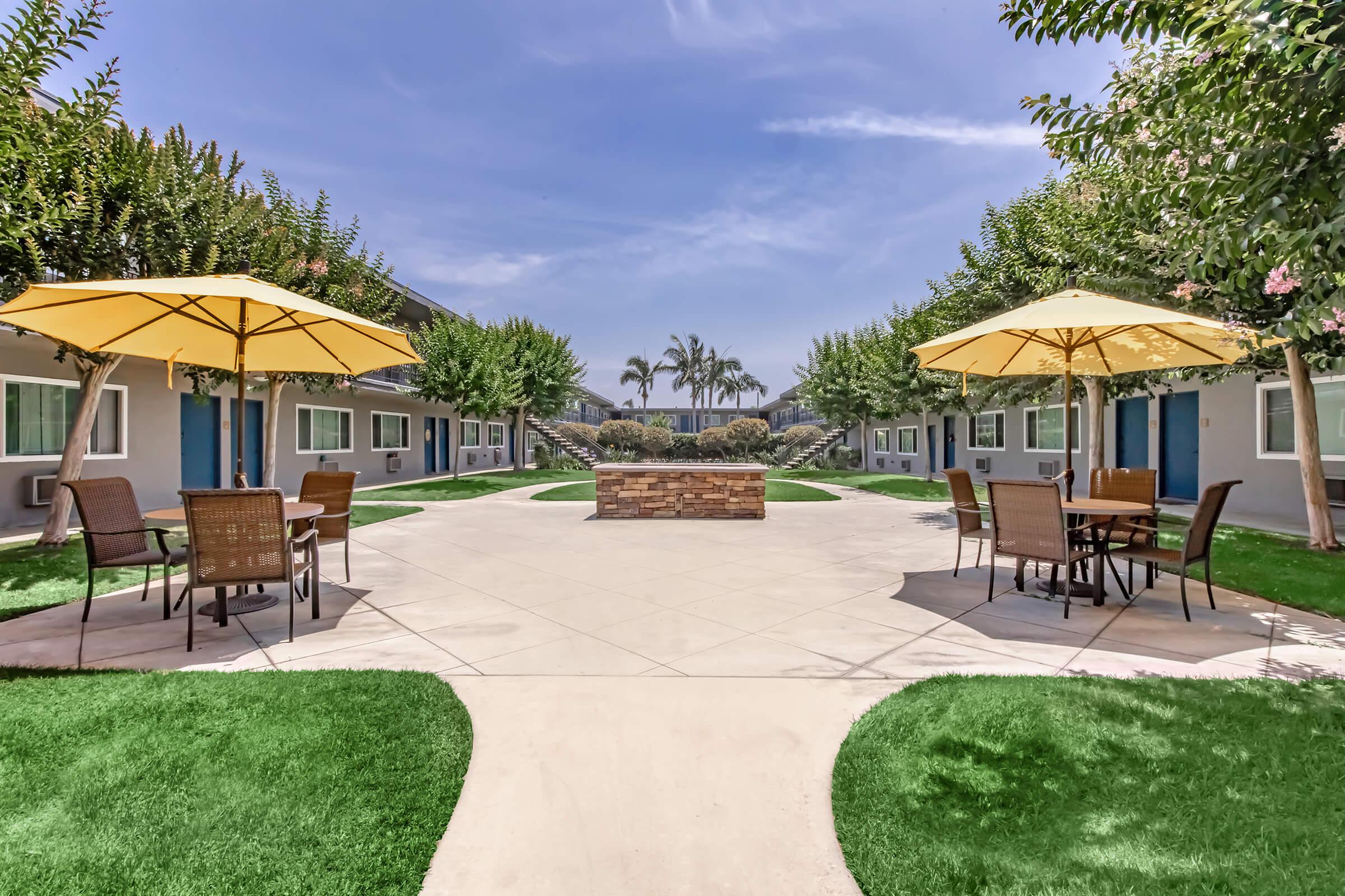 A landscaped courtyard with green grass, two yellow umbrellas over outdoor seating areas, and a stone fire pit in the center. Surrounding the courtyard are light blue buildings with dark blue doors, palm trees in the background, and clear blue sky overhead.