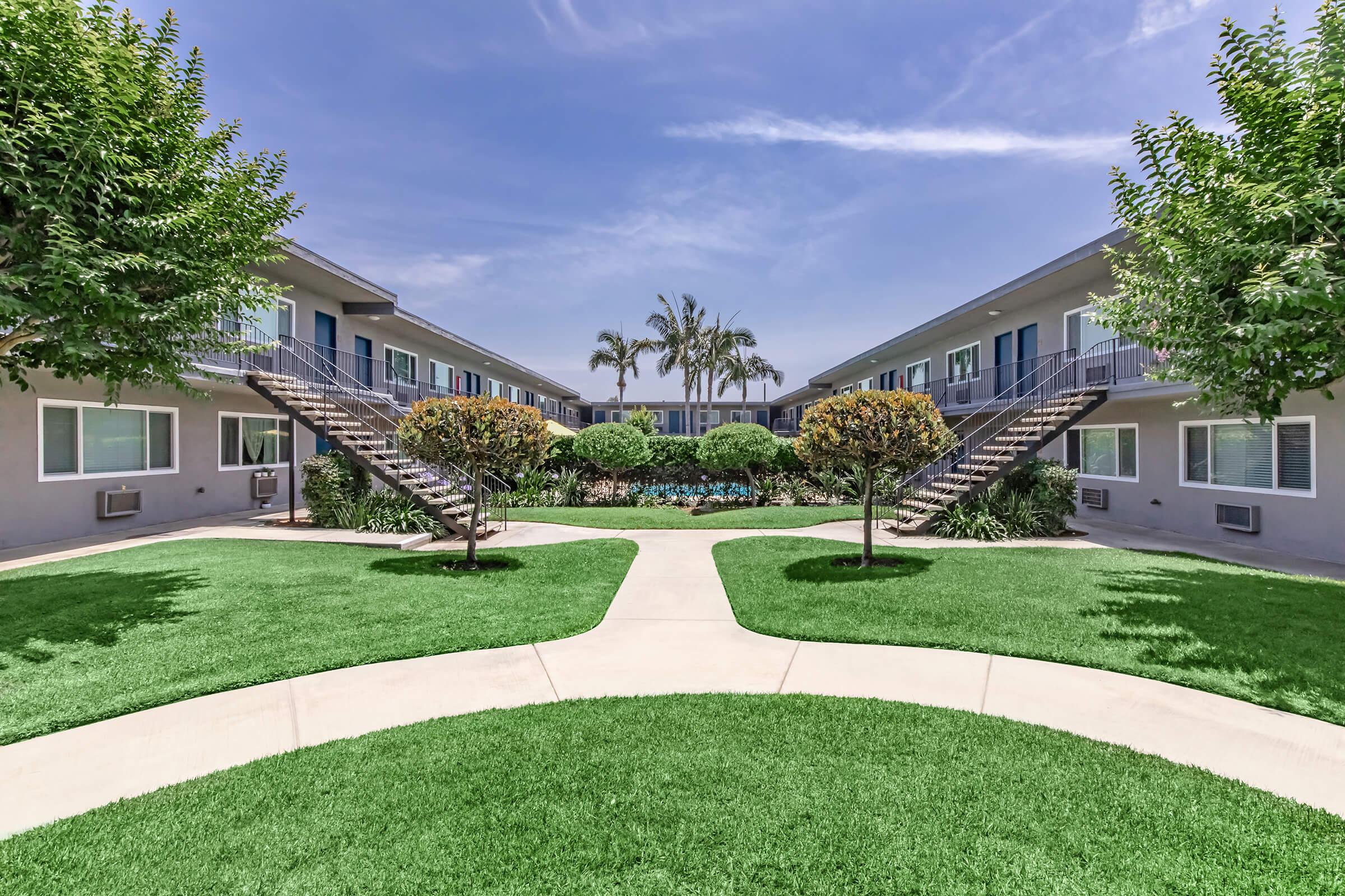A serene courtyard featuring well-maintained green lawns and trees, surrounded by two-story apartment buildings. The pathway diverges in the center, leading to landscaped areas with shrubs and flowers, under a clear blue sky. The setting evokes a peaceful and inviting residential atmosphere.