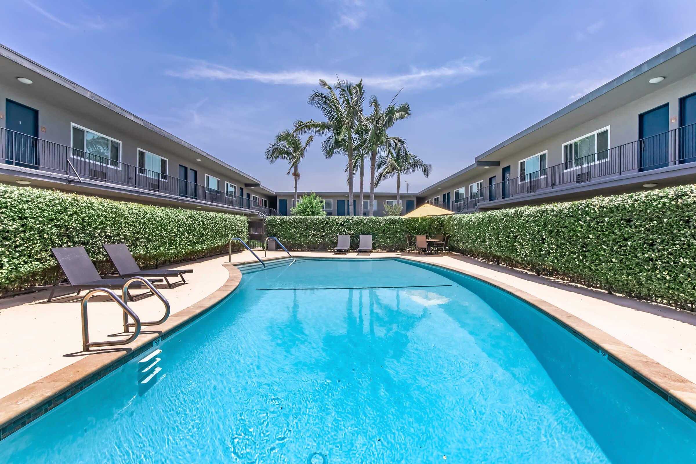 A clear blue swimming pool surrounded by palm trees and green hedges, with sun loungers on the pool deck. In the background, two-story buildings with balconies are visible under a bright blue sky. The scene evokes a relaxing atmosphere perfect for a vacation or leisure.