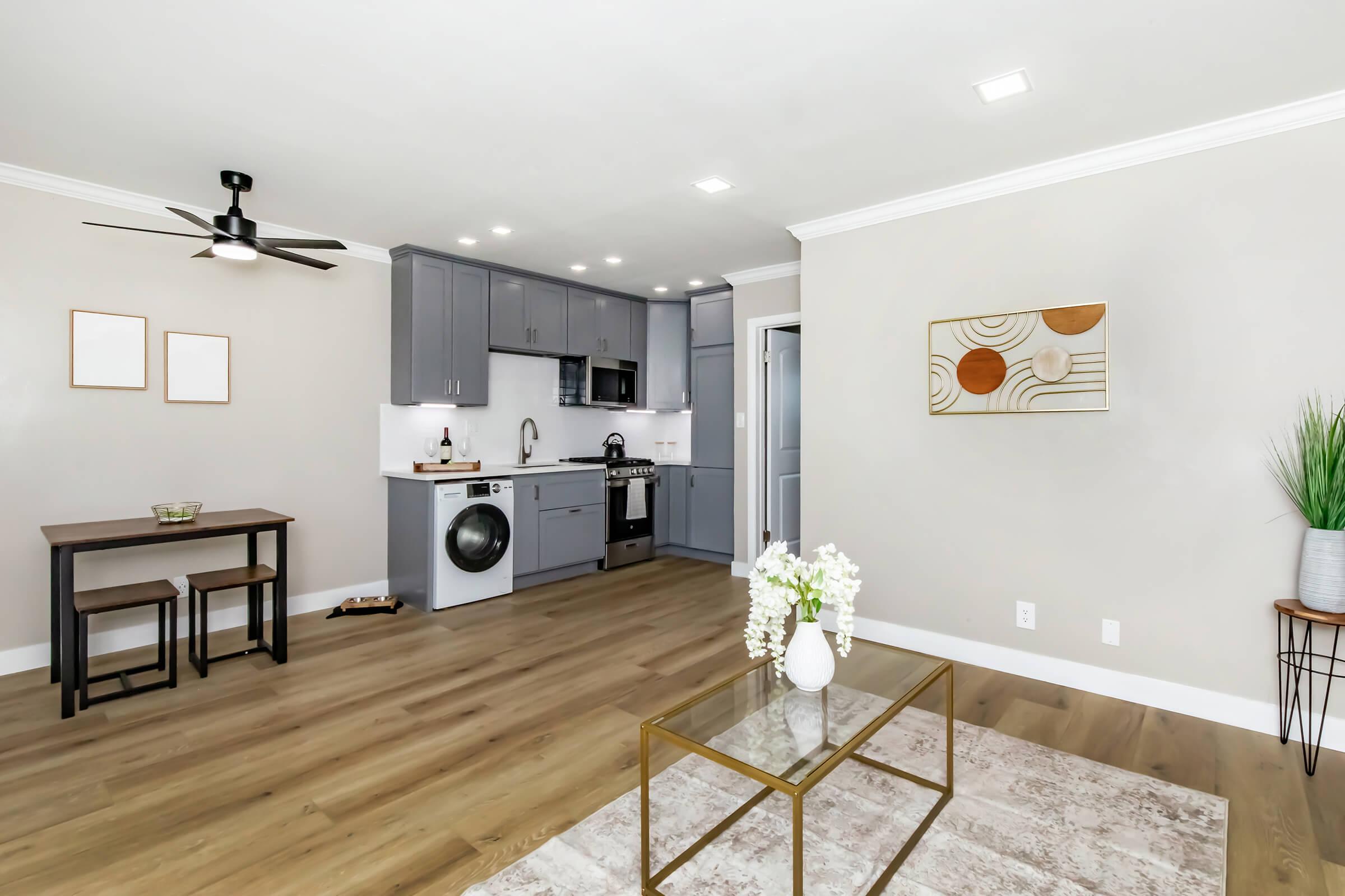 A modern kitchen and living area featuring gray cabinets, stainless steel appliances, a dining table with two chairs, and a coffee table. The space has light-colored walls, wood flooring, and decorative elements like potted plants and wall art, creating a cozy and inviting atmosphere.