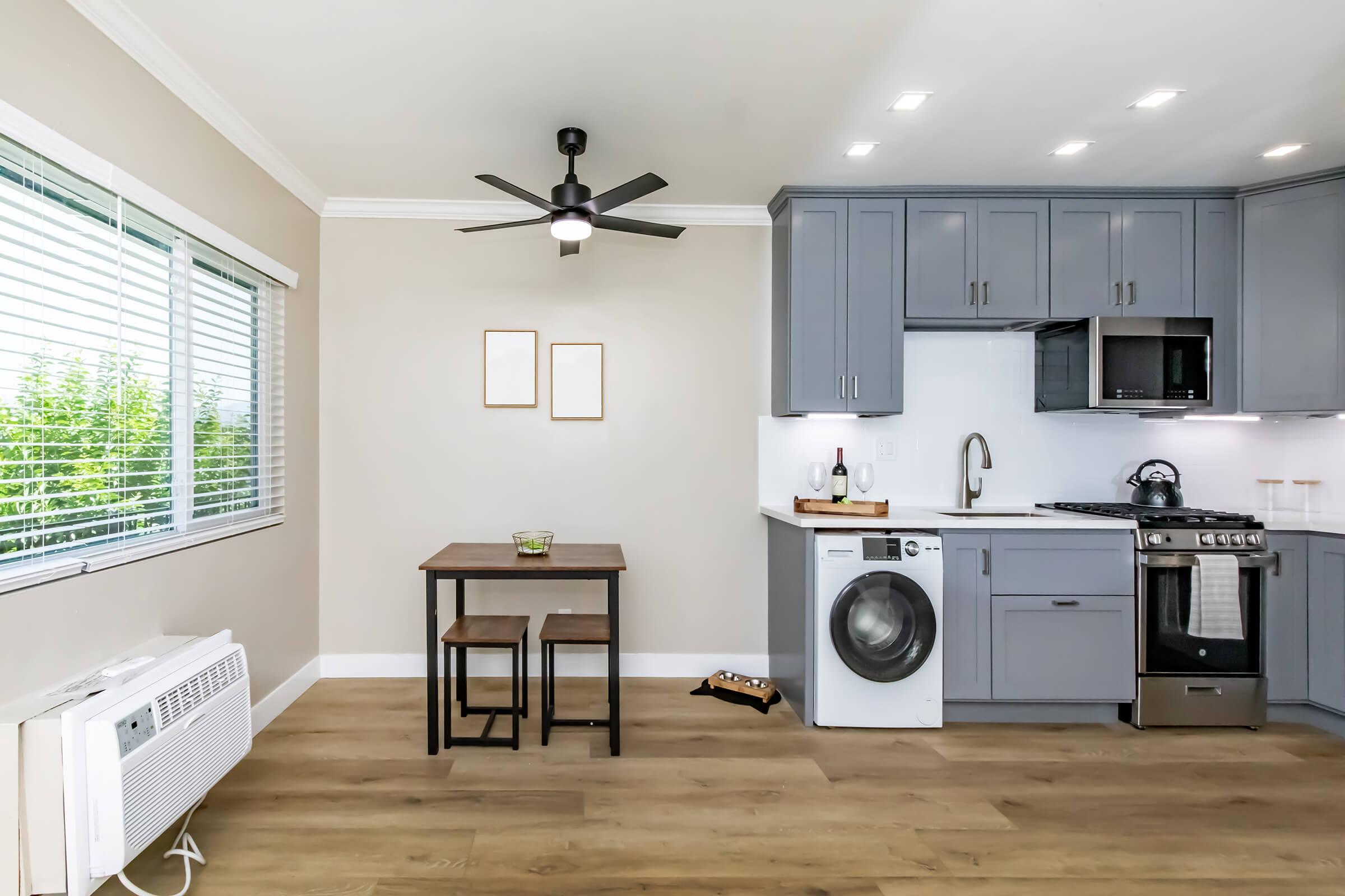 A modern kitchen with gray cabinets, a stainless steel refrigerator, and a washer-dryer combo. A small wooden table with two chairs sits against the wall. Natural light streams in through a window with blinds, and there are two blank framed pictures on the wall. A ceiling fan is visible.