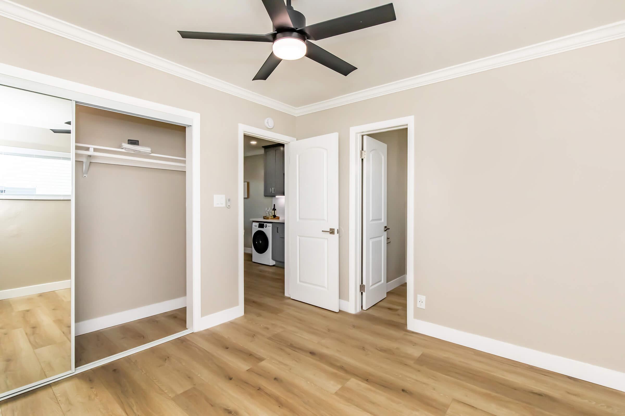 A well-lit interior room featuring light-colored walls, a mirrored closet on the left, and an open doorway leading to another room. The floor has light wood-like laminate, and a ceiling fan is visible above. The room appears spacious and modern, with laundry appliances visible through the doorway on the right.