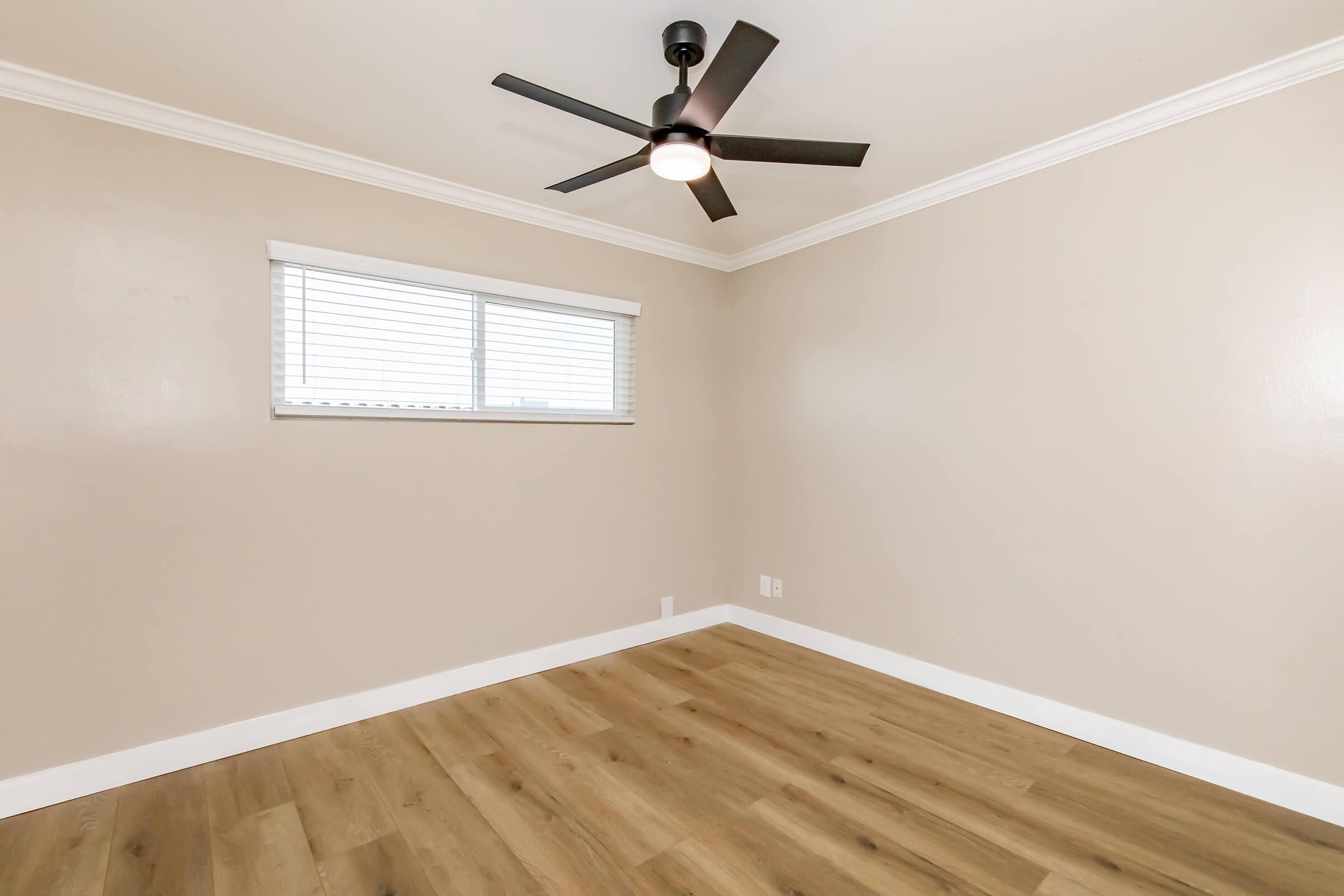 Empty room with light-colored walls, a wooden floor, and a ceiling fan. A window with horizontal blinds allows natural light to enter. The space is uncluttered, offering a blank canvas for potential interior decoration.