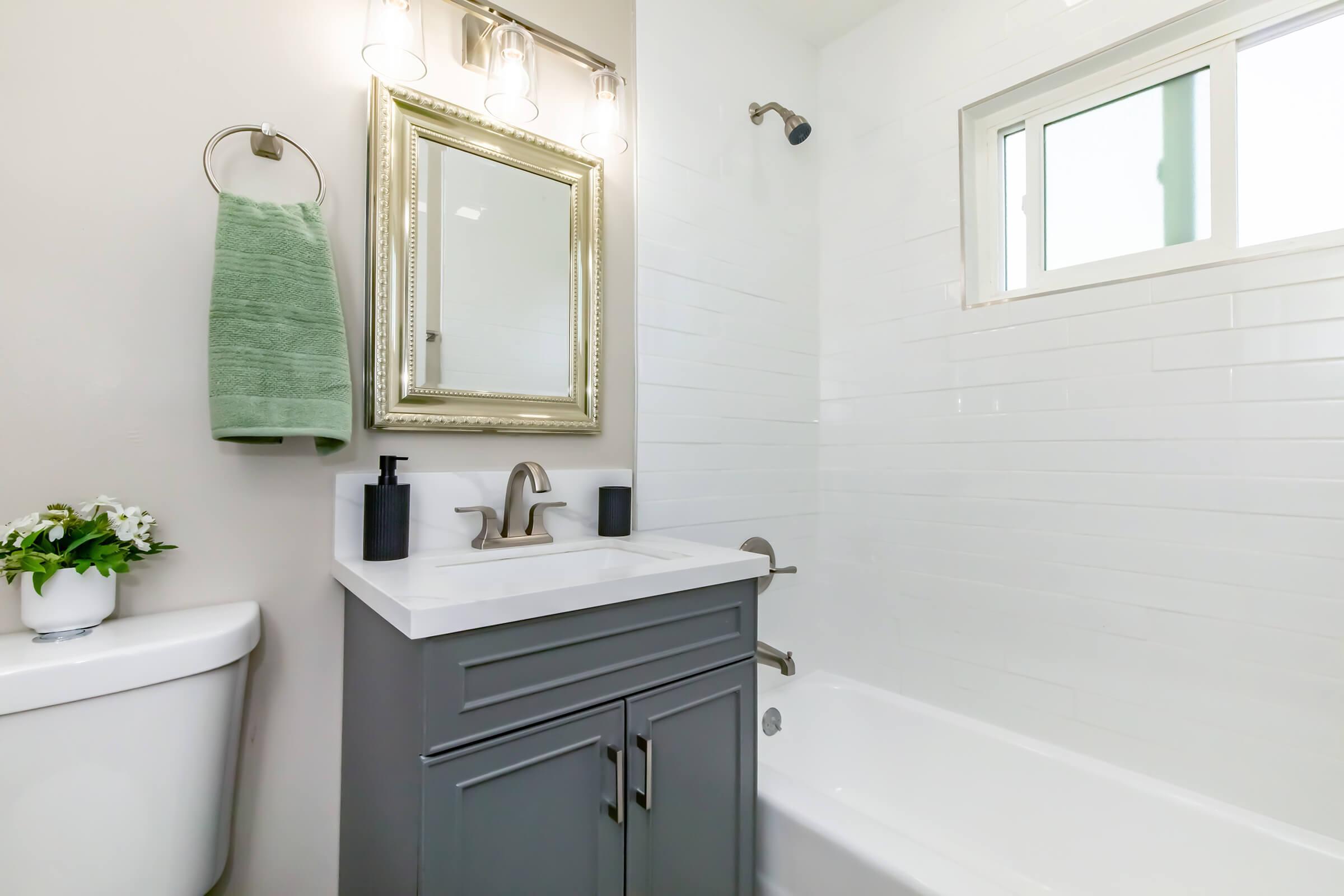 A modern bathroom featuring a bathtub with a shower, a white tiled wall, a gray vanity with a white countertop, a decorative mirror, and a green towel hanging on a towel rack. A small plant is placed on the vanity, and there is a window allowing natural light.
