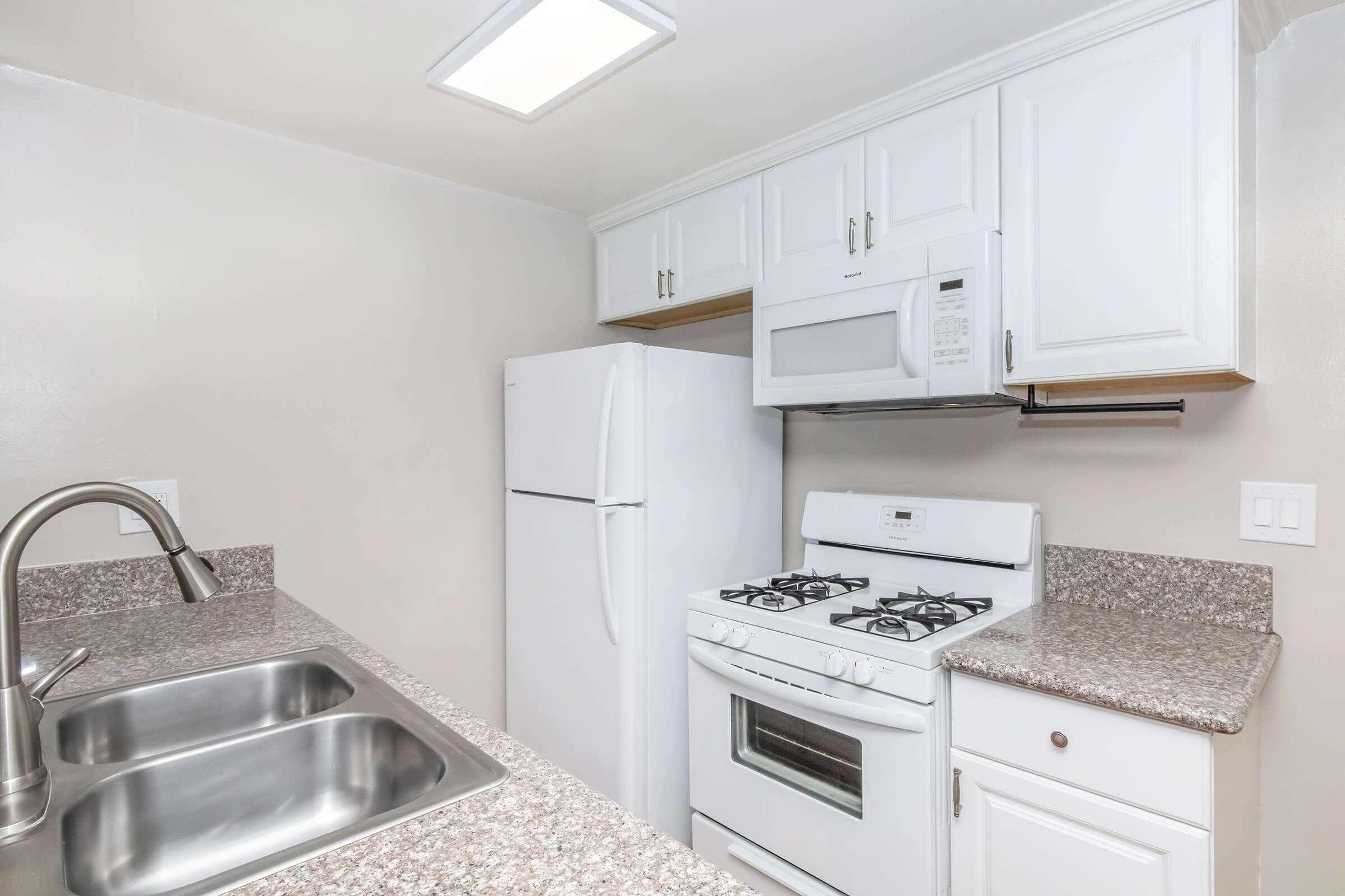 A modern kitchen featuring white cabinetry, a double sink, a gas stove, a microwave, and a white refrigerator. The countertops are made of speckled granite, and there is ample light from a ceiling fixture, creating a bright and inviting space.