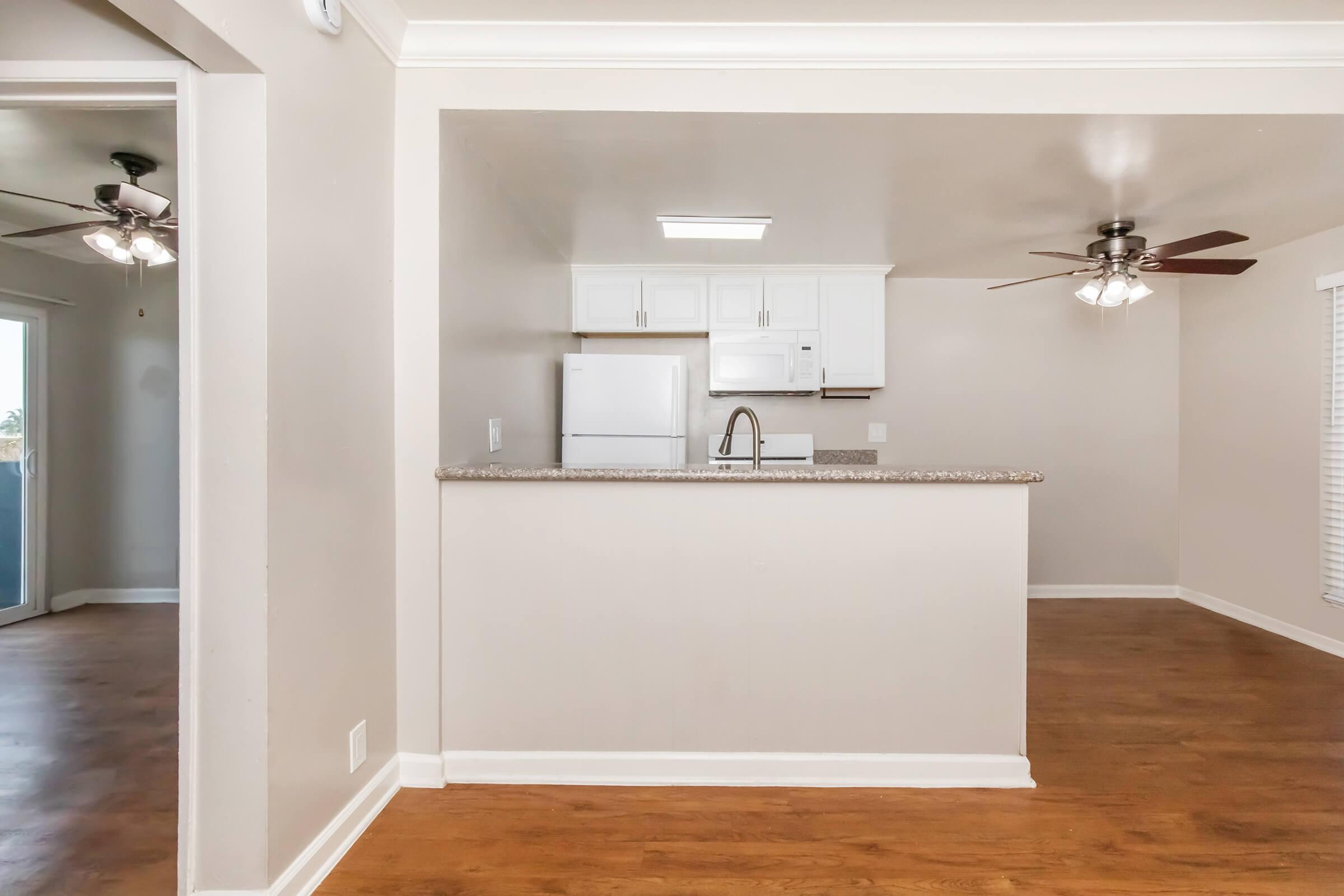 Interior view of a modern kitchen and living area featuring a breakfast bar with a speckled countertop, white cabinets, and a stainless steel sink. Natural light fills the space, highlighting the wooden floor. Two ceiling fans are visible, and a doorway leads to another room.