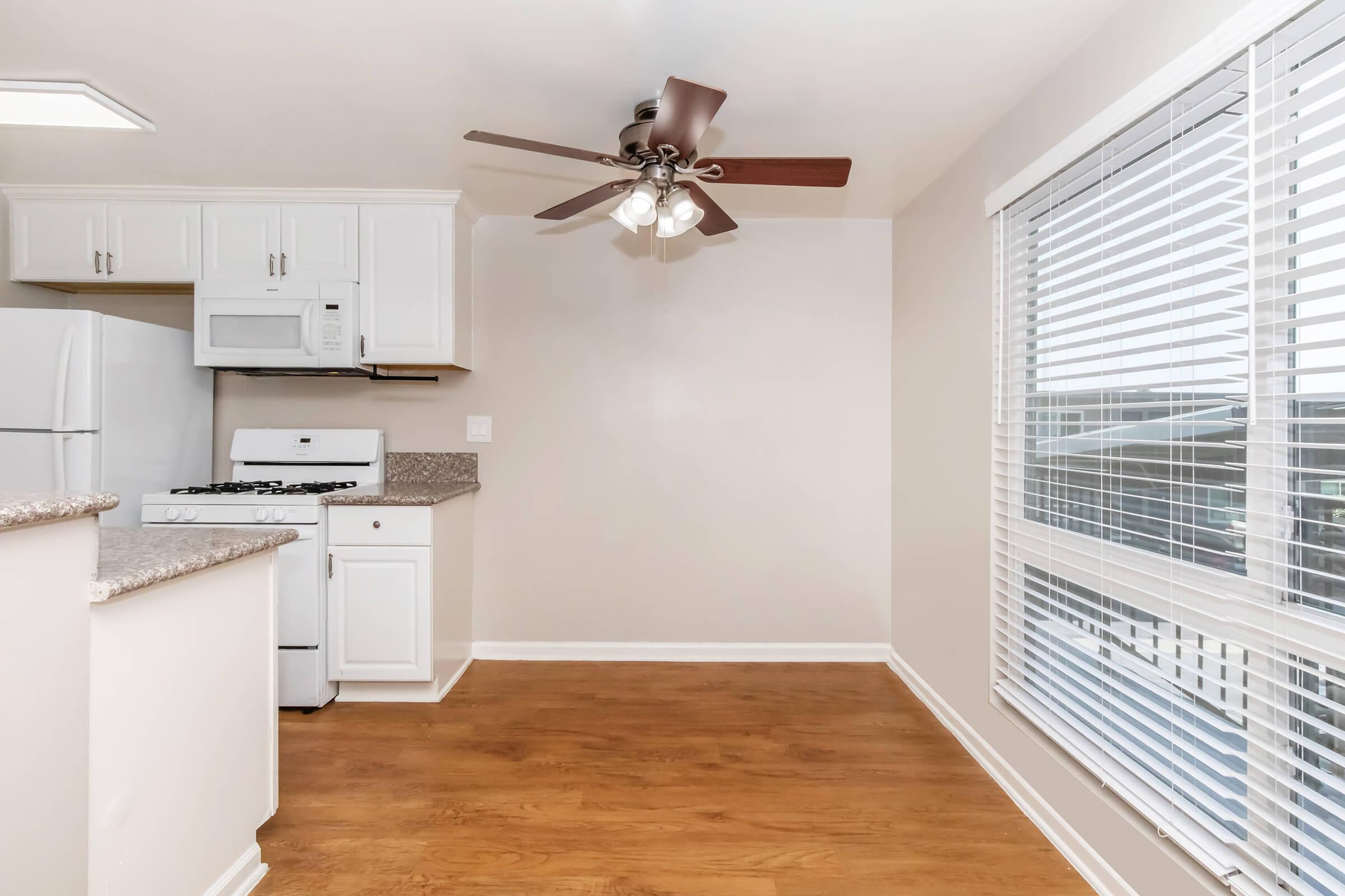 Bright, modern kitchen with white cabinets, a white refrigerator, and a gas stove. Features a granite countertop and a ceiling fan with lights. Large window with blinds allows natural light, highlighting the warm wood floor. The layout is open, creating a spacious feel.