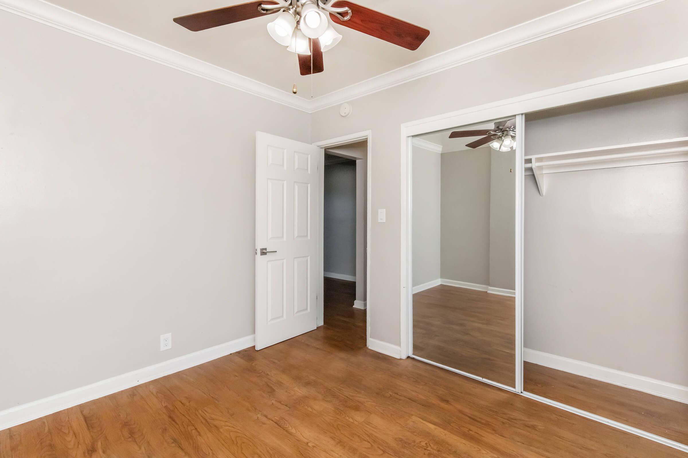A well-lit room featuring light gray walls and a ceiling fan with five blades. There is a white door leading to another space, and a mirrored closet door showcasing a wooden floor. The room is empty, creating a clean and spacious feel.