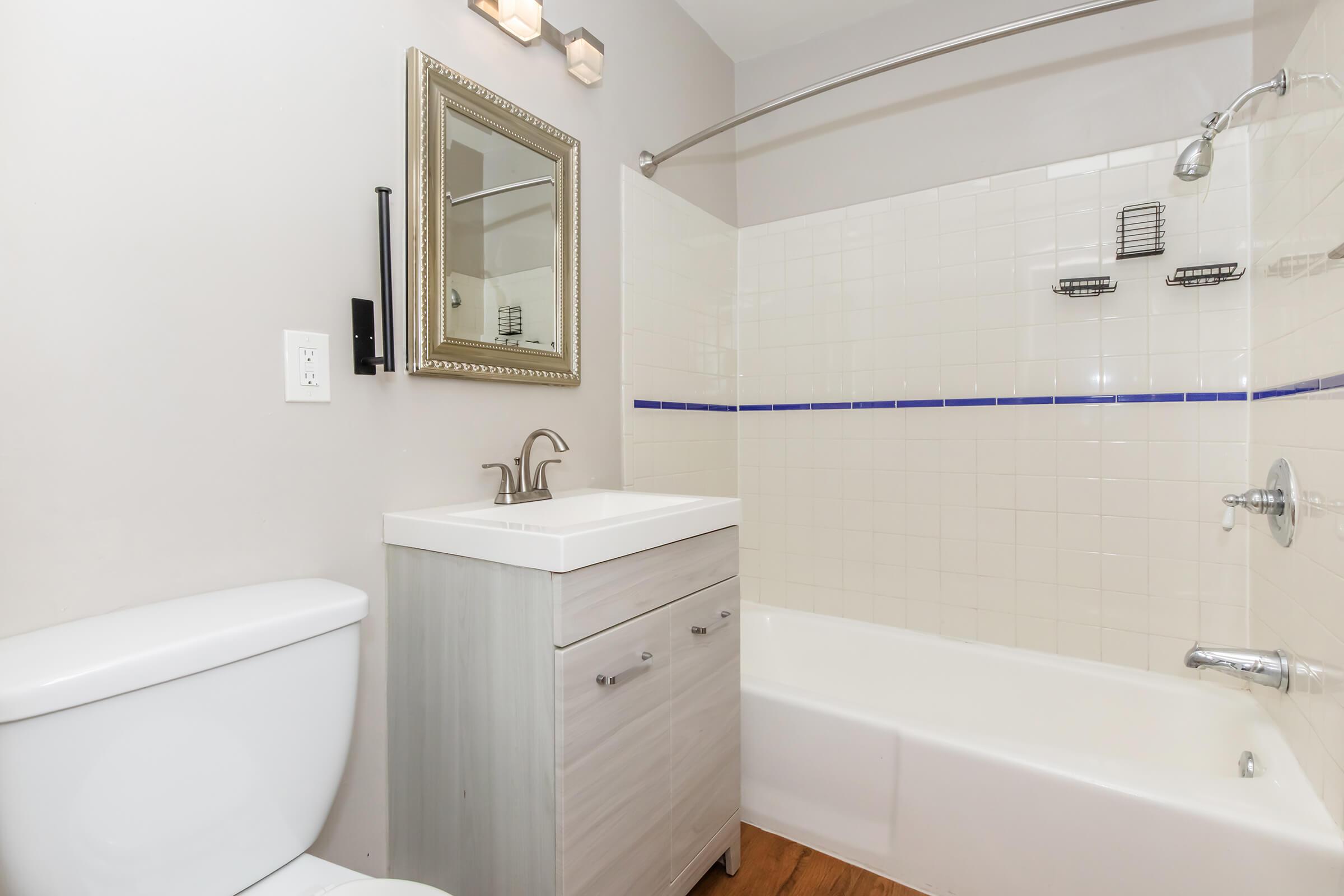 A modern bathroom featuring a white toilet, a sleek vanity with a sink, and a bathtub with a shower. The walls are light-colored with a decorative mirror above the vanity. The tub area has white tiles with a blue accent stripe. The flooring is a warm wood tone, creating a contemporary look.