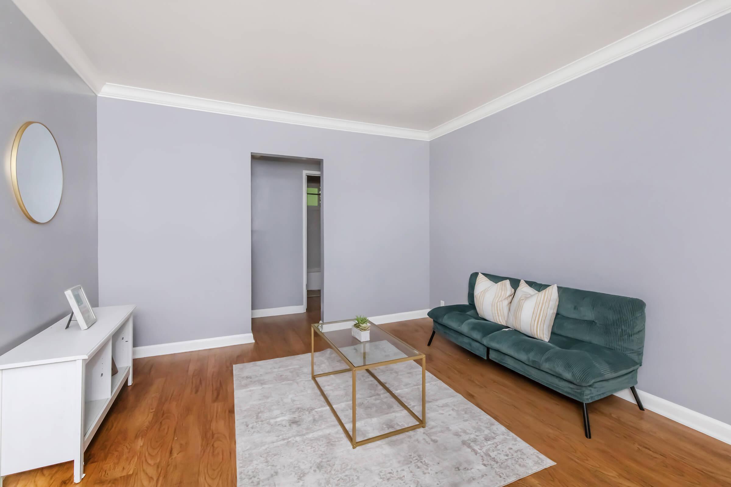 A modern living room featuring a light purple wall, a stylish green sofa with decorative pillows, a glass coffee table with a small plant, and a white media unit. There is a round mirror on the wall and a doorway leading to another room, with hardwood flooring throughout.