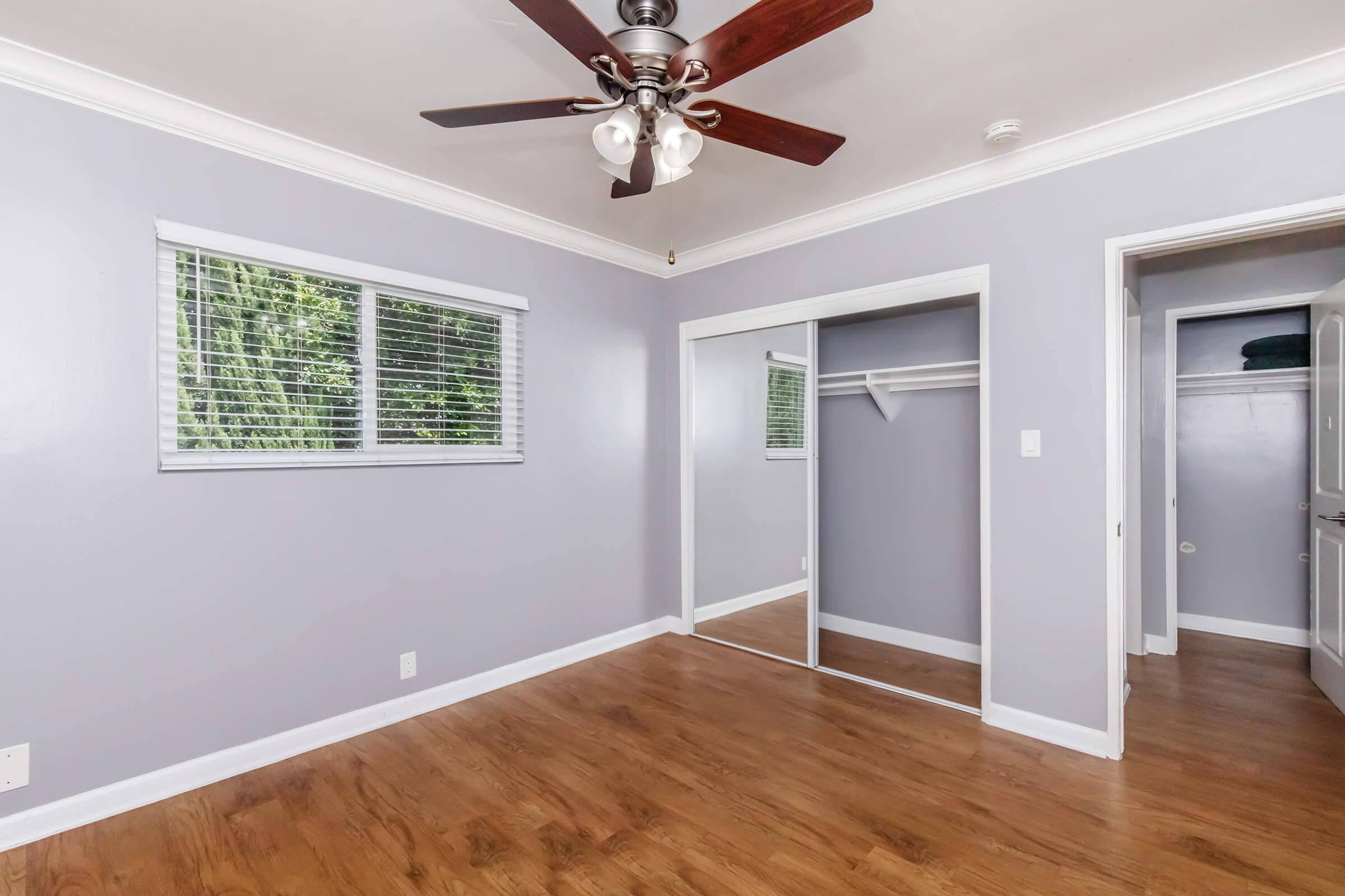 A simple, modern bedroom featuring light gray walls, a ceiling fan, and hardwood flooring. There is a window with blinds allowing natural light, a mirrored closet door, and an open doorway leading to another space. The room is clean and uncluttered, creating a bright and inviting atmosphere.