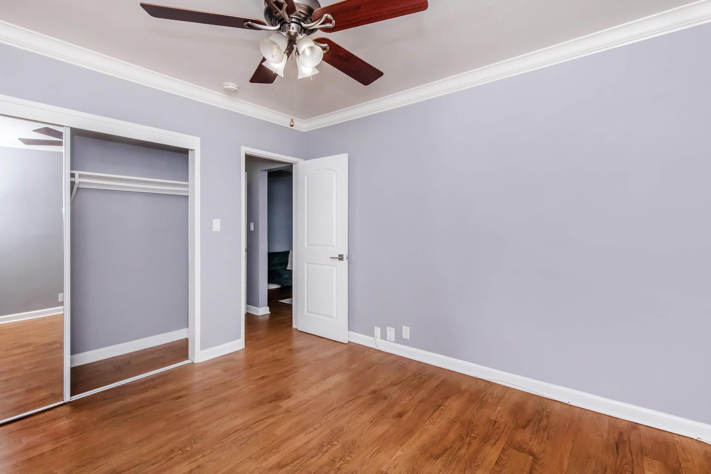 A well-lit room with light purple walls and a wooden floor. It features a ceiling fan, a closet with mirrored doors, and a doorway leading to another room. The overall atmosphere is clean and inviting, suitable for a bedroom or office space.