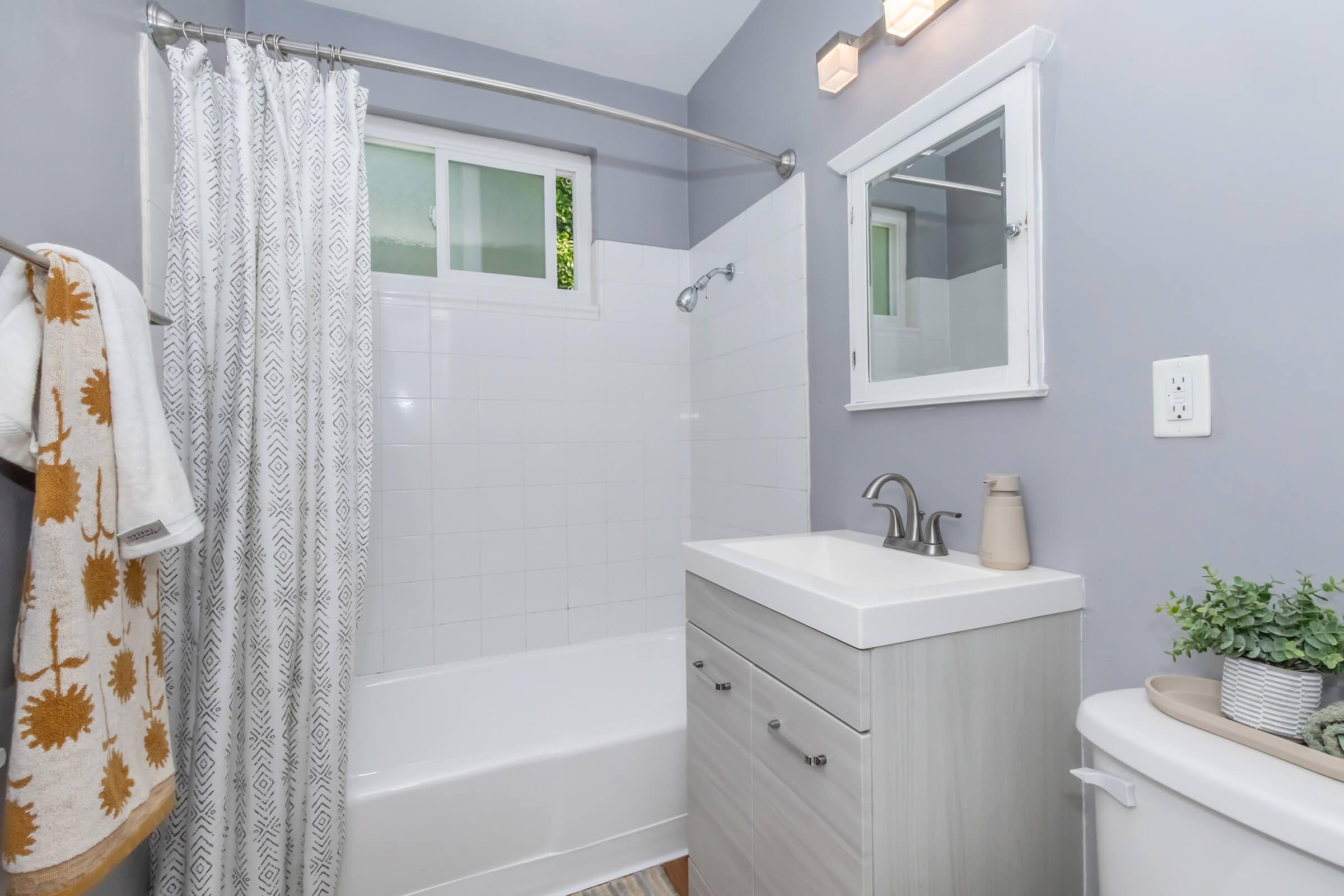 A modern bathroom featuring a white bathtub with a shower curtain, a light gray vanity with a sink, a small mirror, and minimal decorations. Soft natural light comes through a window, and there are decorative elements like a potted plant and towels on a rack. The walls are painted light gray.
