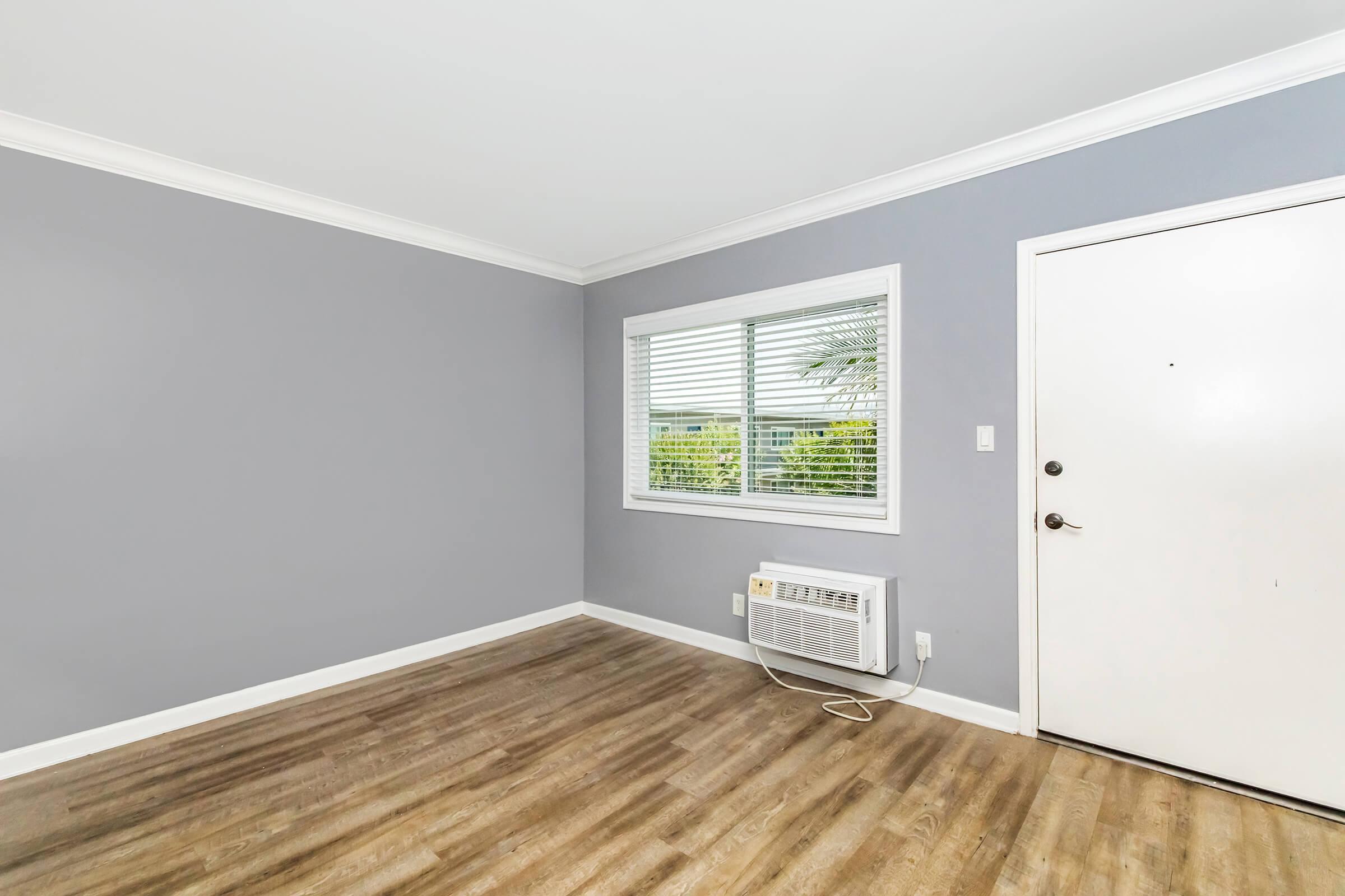 A light gray room with a window featuring white blinds, allowing natural light to enter. There is an air conditioning unit on the floor beneath the window. The flooring is a light wood-like laminate, and the door is white with a round doorknob. The walls are clean and free of decorations.
