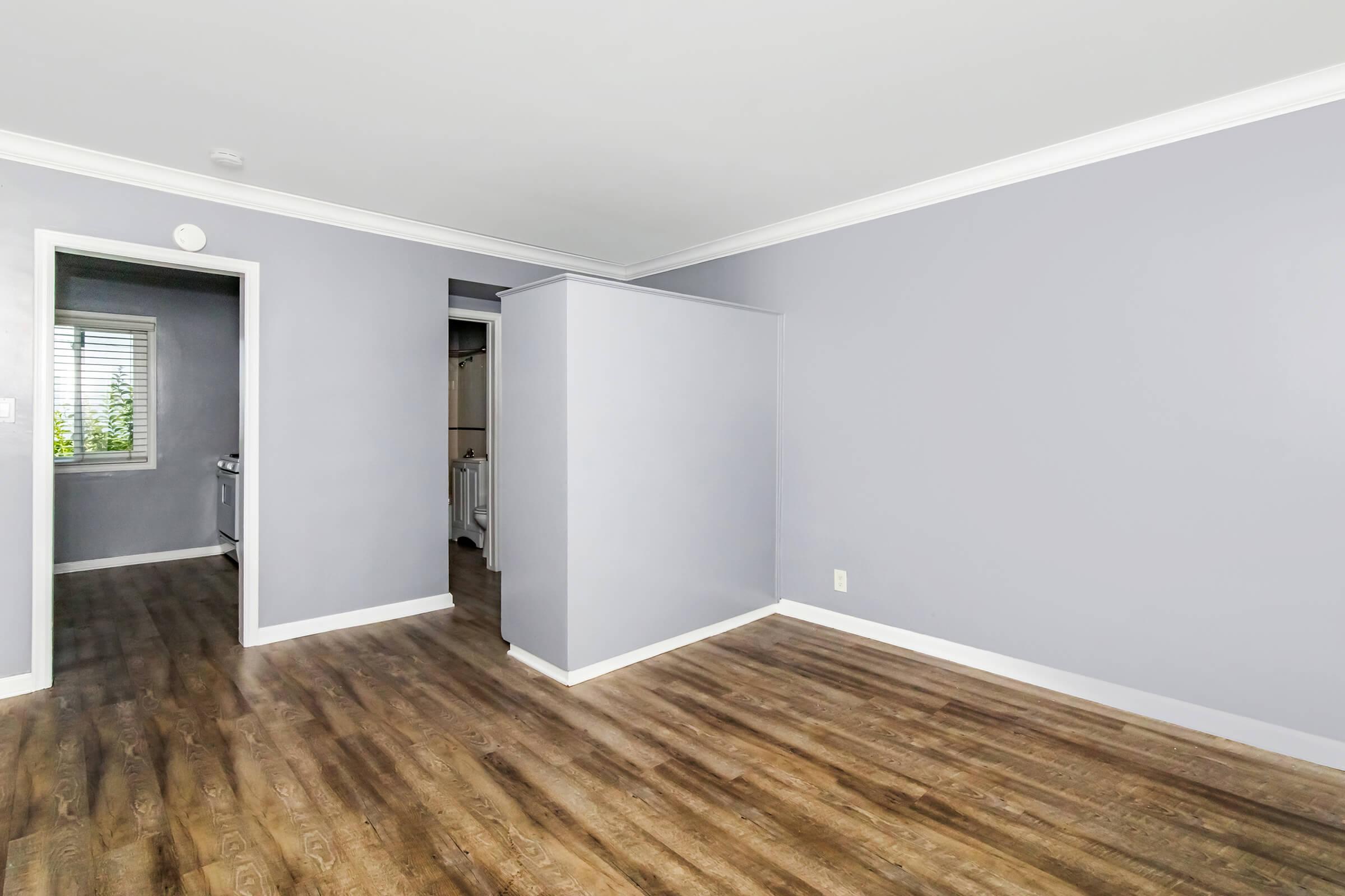 A spacious room with light gray walls and laminate flooring. There is a doorway leading to another room and a window providing natural light. The overall atmosphere is bright and inviting, suitable for various uses.