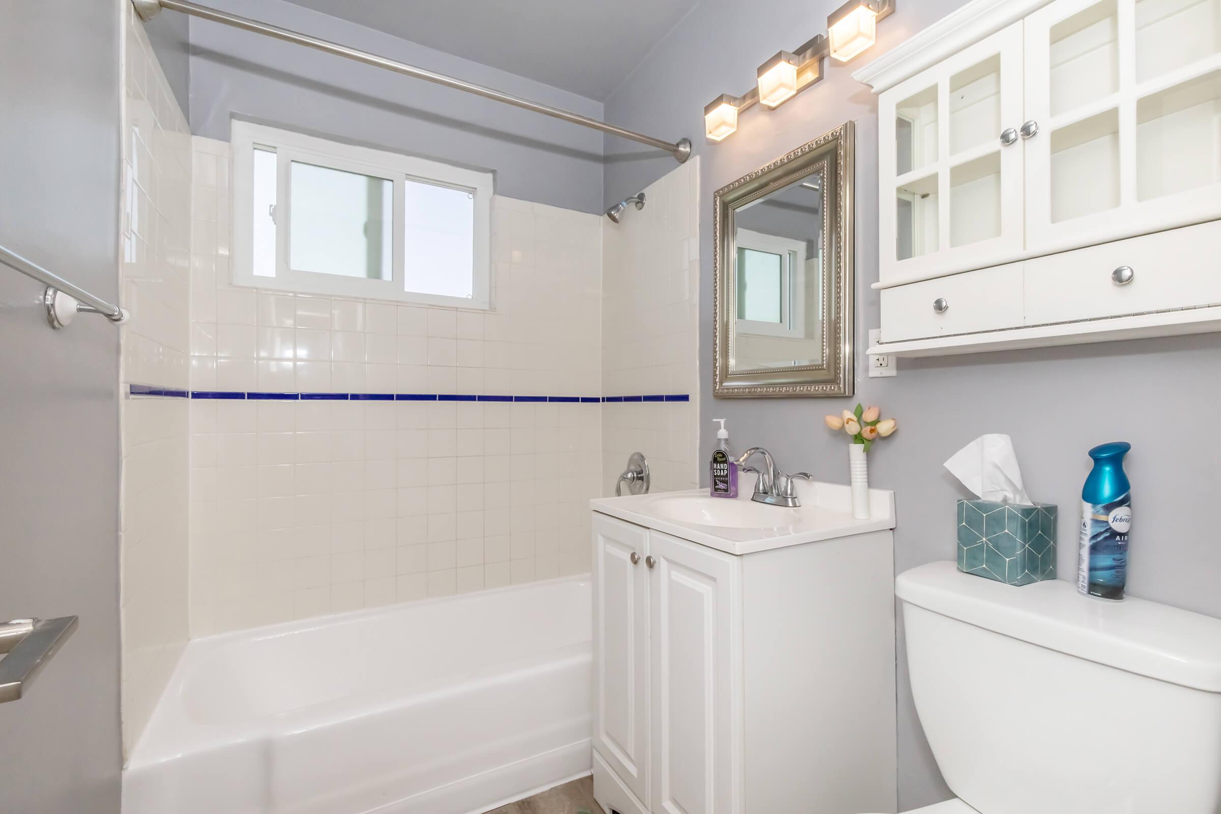 A bright, modern bathroom featuring a white bathtub with a blue accent tile, a white vanity with a sink, a toilet, and a large mirror. Natural light enters through a small window. The decor is simple and clean, with a few toiletries and a small flower arrangement on the vanity.