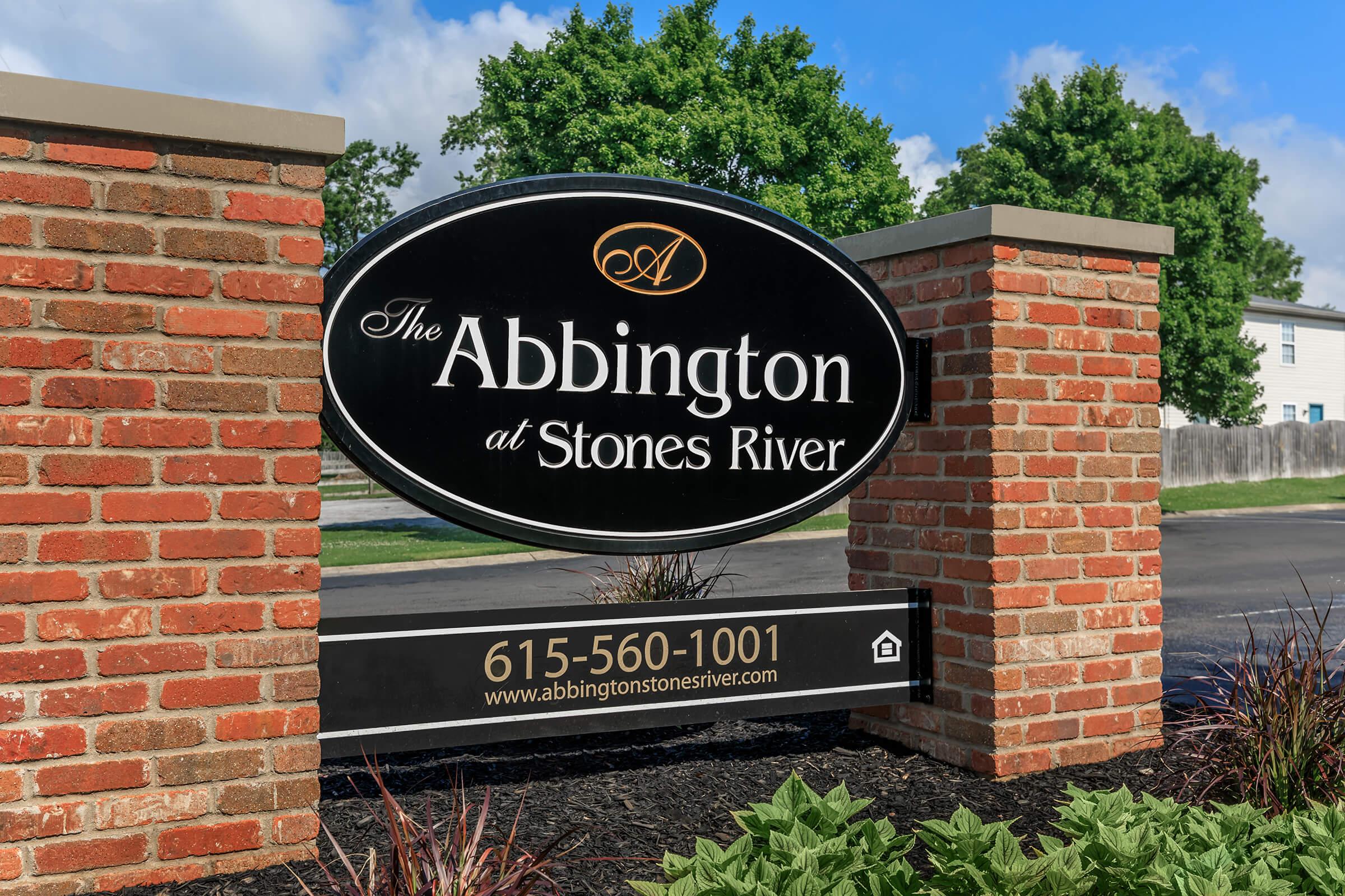 A black oval sign with the text "The Abbington at Stones River" in white letters, surrounded by a red brick structure. Below the sign, there's a phone number "615-560-1001" and a website address "www.abbingtonstonesriver.com." Green trees and a blue sky are visible in the background.