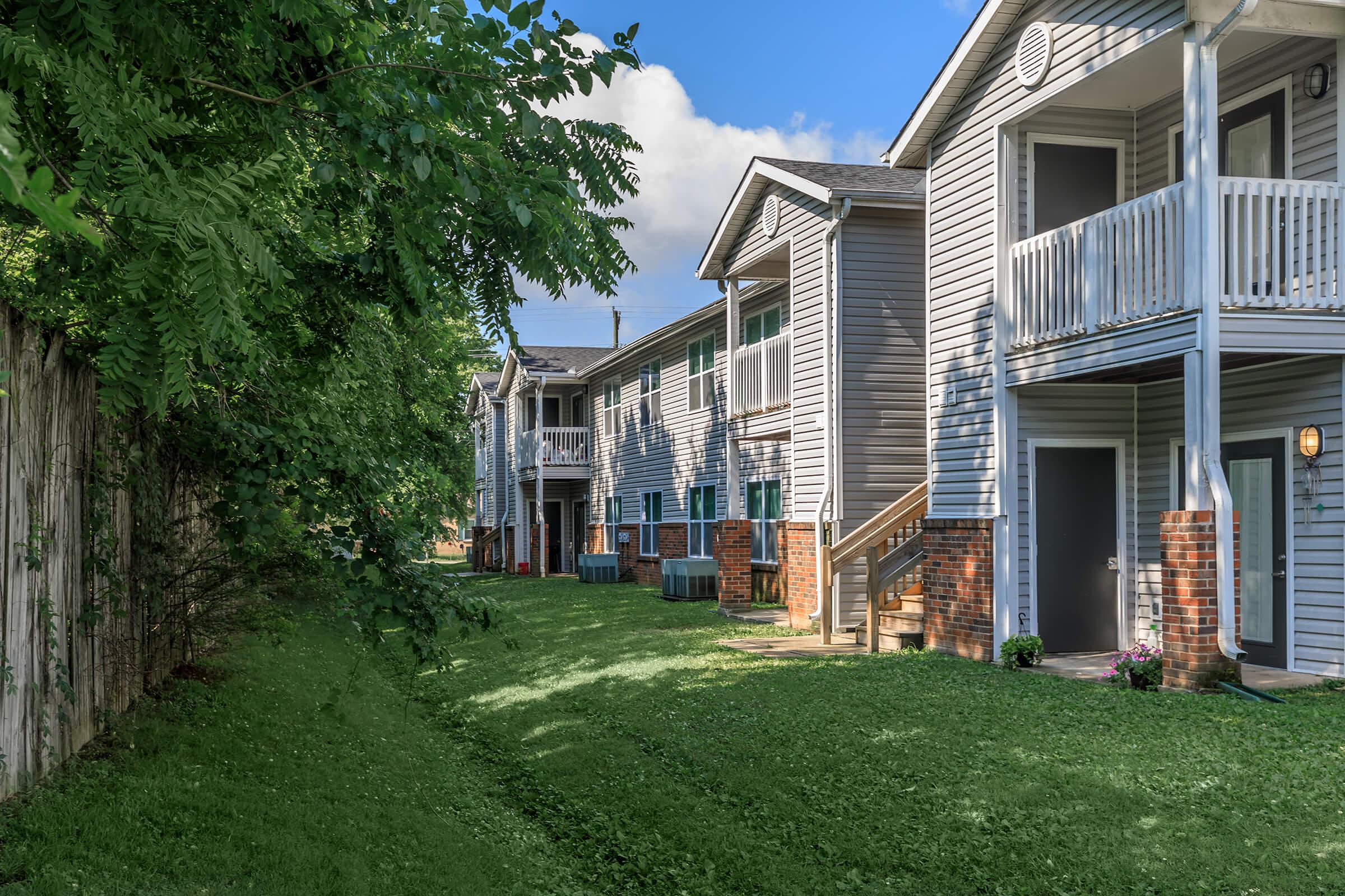 A row of apartment buildings with balconies, surrounded by a green lawn and trees. The buildings feature grey siding and brick accents, with a clear blue sky above. The scene conveys a peaceful residential atmosphere.