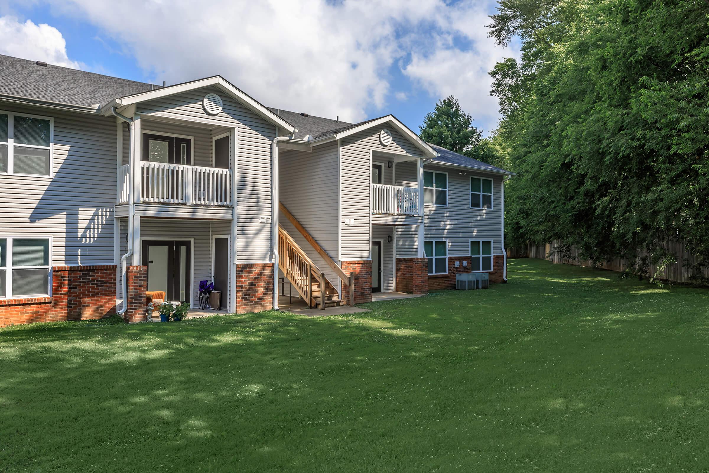 Two-story residential building with light gray siding and white trim. The structure features balconies on the upper level and a set of stairs leading to the ground level. A well-manicured green lawn surrounds the building, with some trees in the background and scattered outdoor furniture on the patio area.