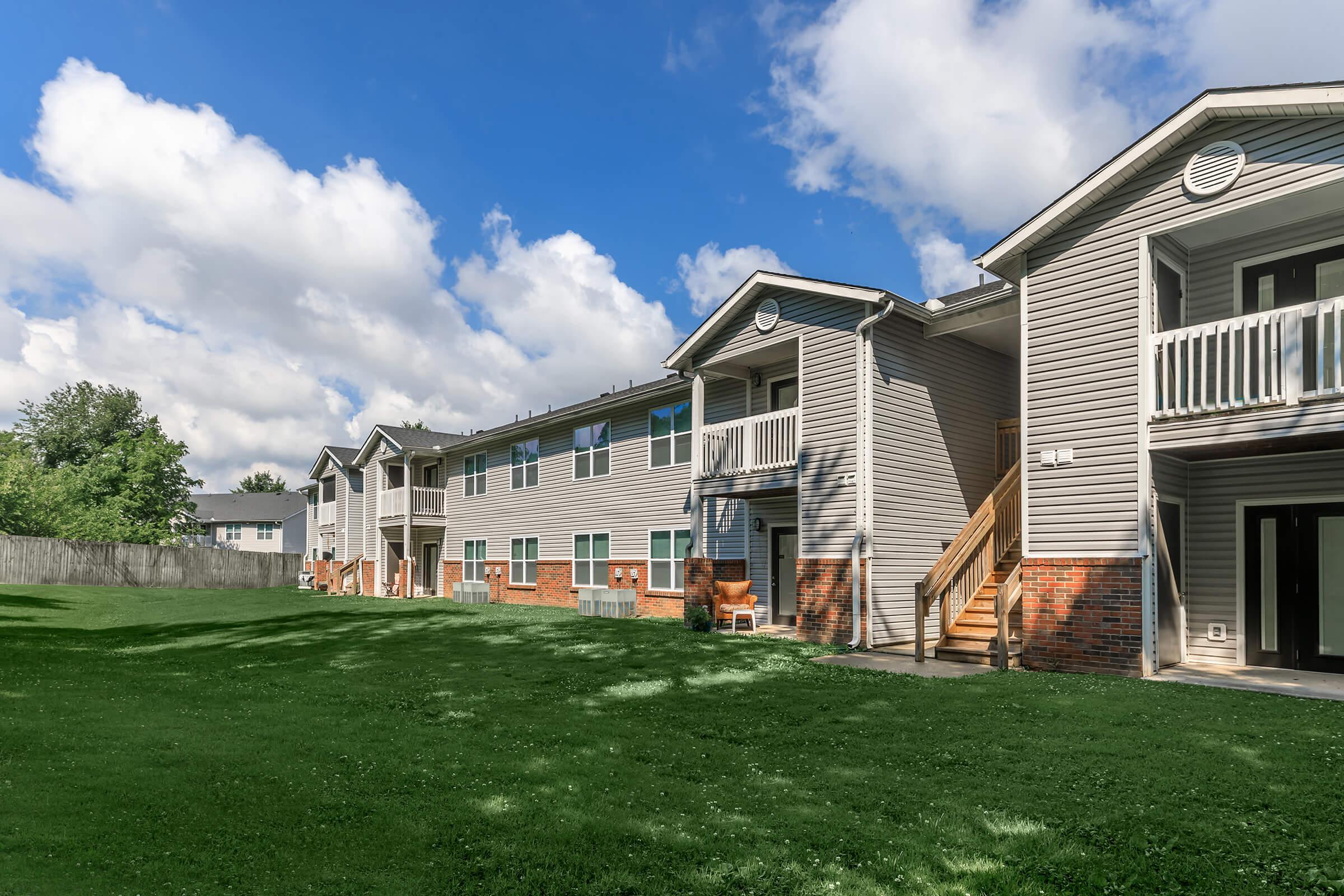 Apartment buildings with multiple balconies are set against a vibrant blue sky with scattered clouds. Lush green grass covers the foreground, and a wooden fence borders the property, providing a serene residential atmosphere. The buildings feature a mix of siding and brick exteriors.