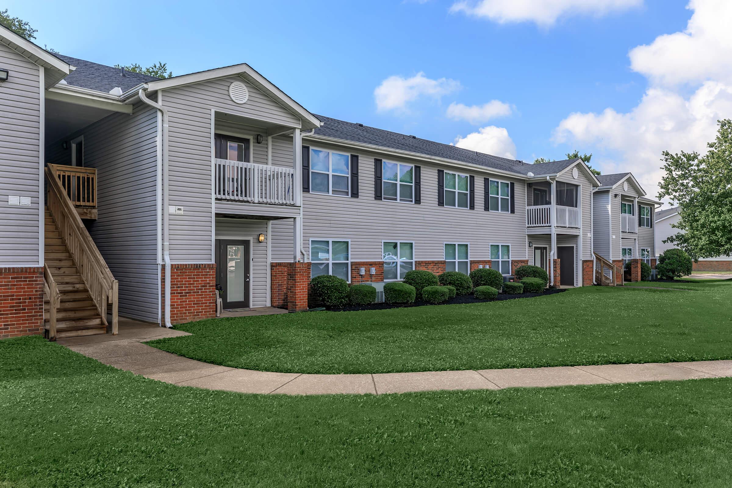 A well-maintained apartment complex with a two-story building featuring a mix of balconies and windows. The exterior is light gray with brick accents, surrounded by a neatly landscaped lawn and pathways. The sky is partly cloudy, contributing to a pleasant residential atmosphere.