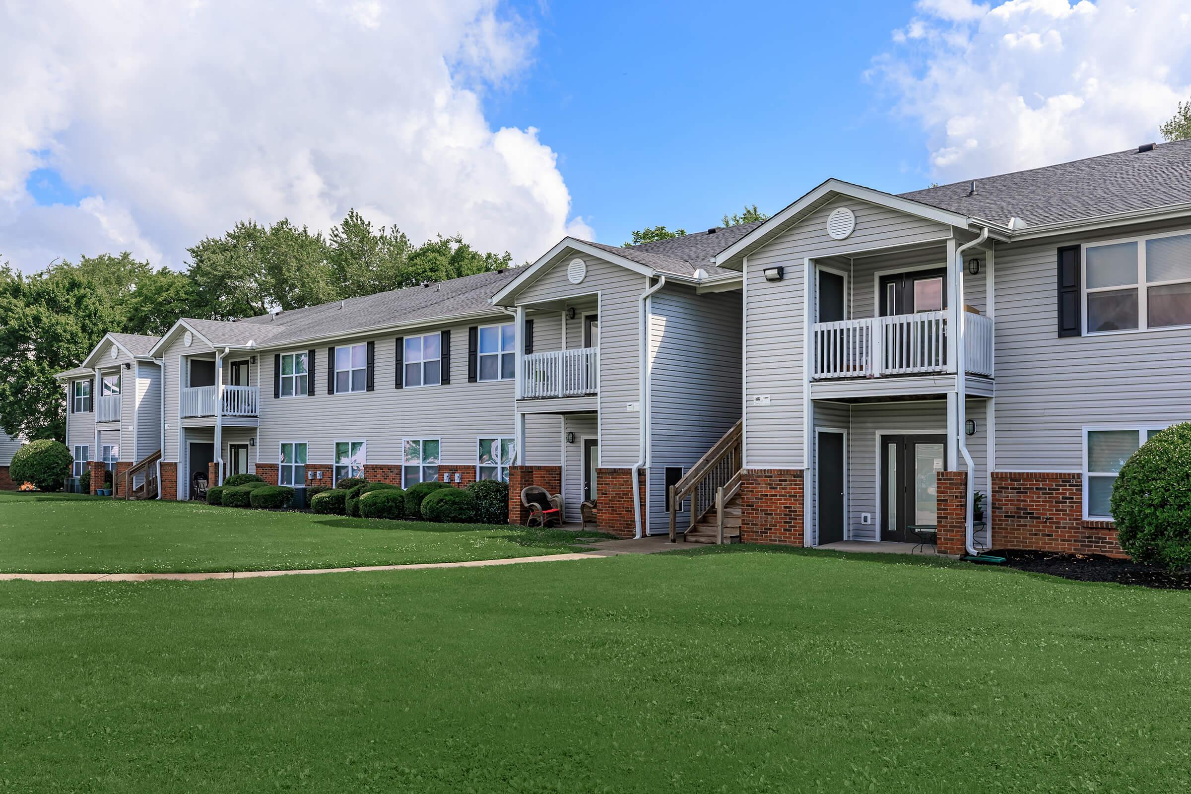 A row of multi-unit apartment buildings with light gray siding and red brick accents. Each unit has a small balcony and stairs leading up to the entrances. The landscaping features a manicured lawn and shrubs, set against a backdrop of blue skies with white clouds.
