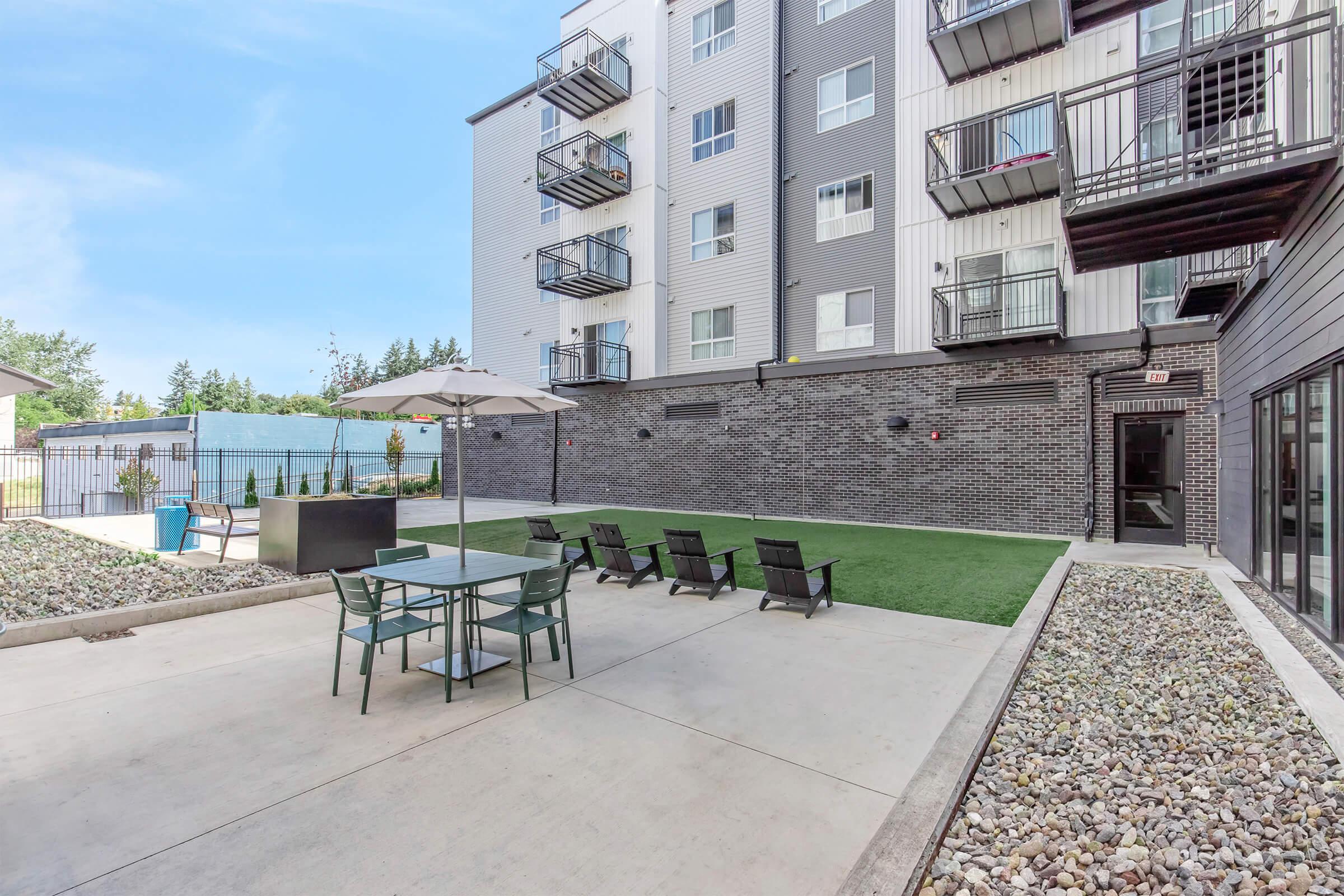 Outdoor area of an apartment complex featuring a landscaped lawn, a patio table with chairs, lounge chairs, and an umbrella. In the background, there are multiple apartment balconies and a fenced area with greenery. The setting is bright and well-maintained, ideal for relaxation or social gatherings.