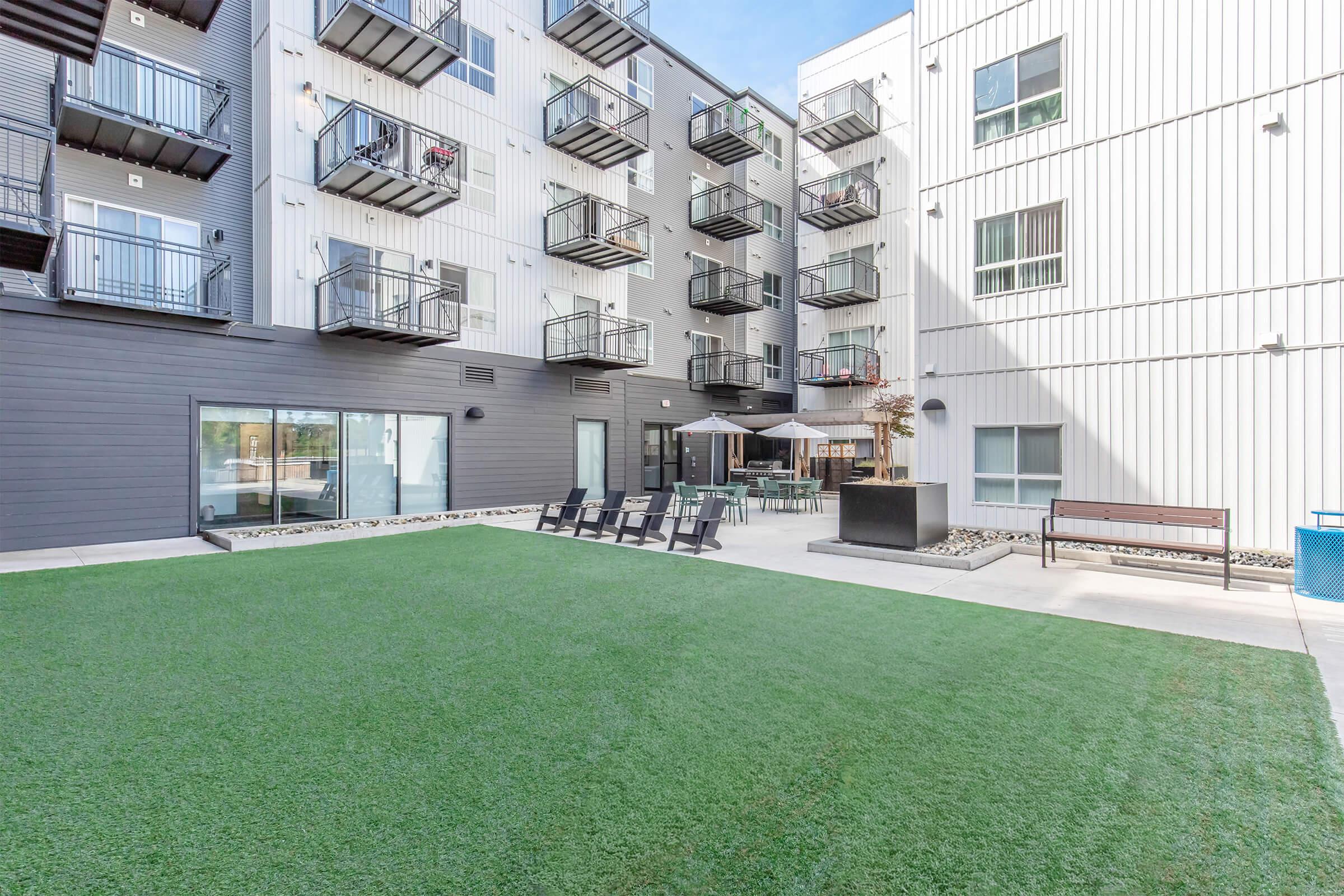 A modern apartment complex courtyard featuring green artificial grass, black lounge chairs, and multiple balconies. There are tables and umbrellas for outdoor seating, with a large glass door leading into the building. The exterior walls are a mix of dark and light materials, creating a contemporary look.