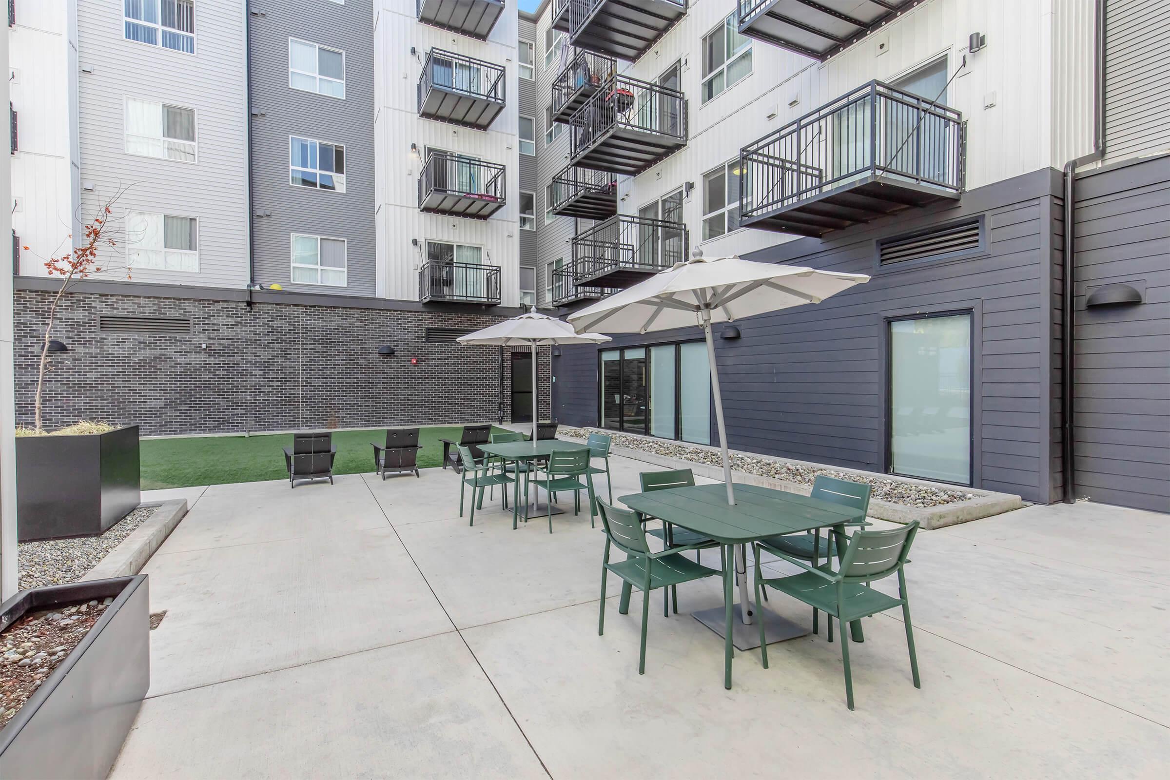 Outdoor courtyard area with green tables and chairs under white umbrellas. Surrounding walls are modern with balconies visible above. A grassy area is seen in the background, along with decorative planters. The overall atmosphere is inviting and suitable for relaxation or social gatherings.