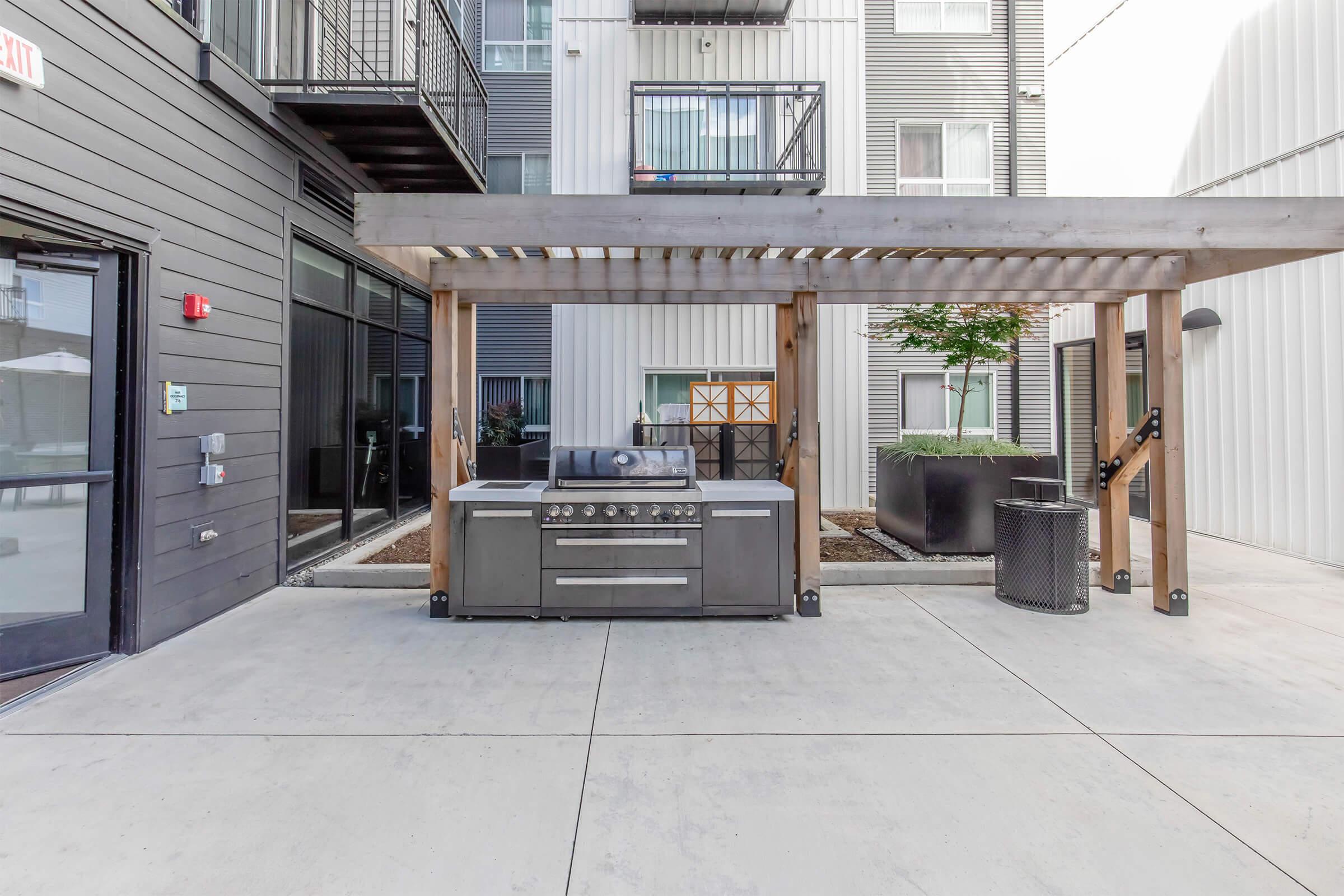 An outdoor grilling area featuring a modern barbecue grill under a wooden pergola. The backdrop includes a multi-story building with large windows, and there are planters with greenery. The area is paved with concrete and has a contemporary, clean design.