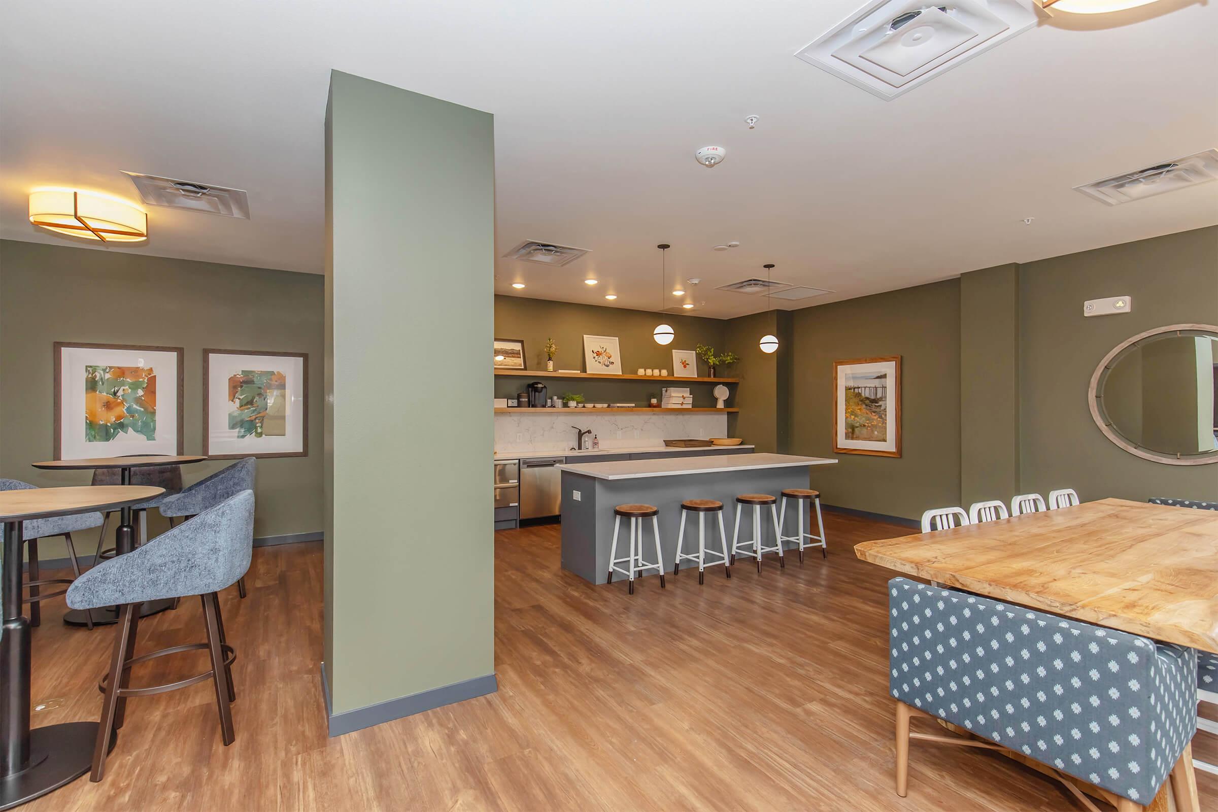 Modern common area featuring a kitchenette with gray cabinetry and bar stools, a dining area with a wooden table and chairs, and artwork on the walls. The space has green accents and hardwood flooring, creating a welcoming atmosphere for relaxation and socializing.