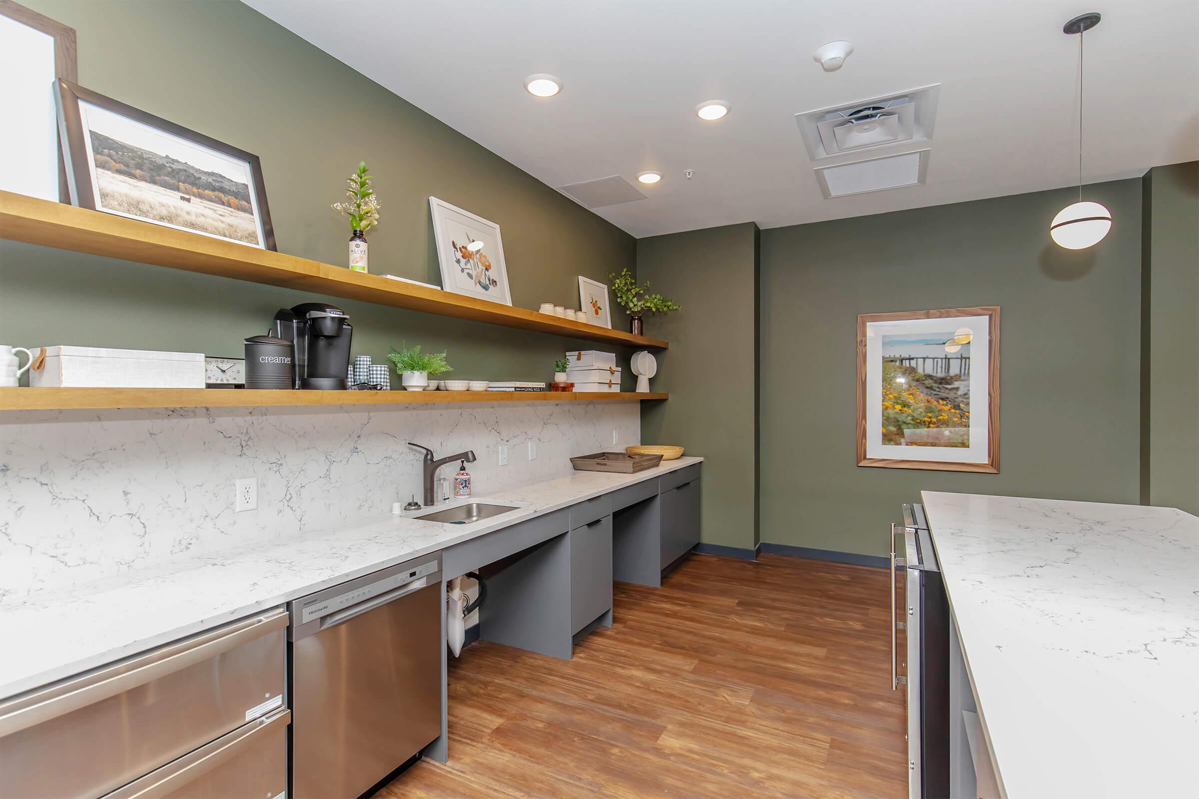 A modern kitchen featuring a marble countertop, stainless steel sink, and dishwasher. Open wooden shelves display decorative items and kitchen essentials. The walls are painted a muted green, and there is a large window with a framed picture of a landscape decoratively placed. The flooring is wood.