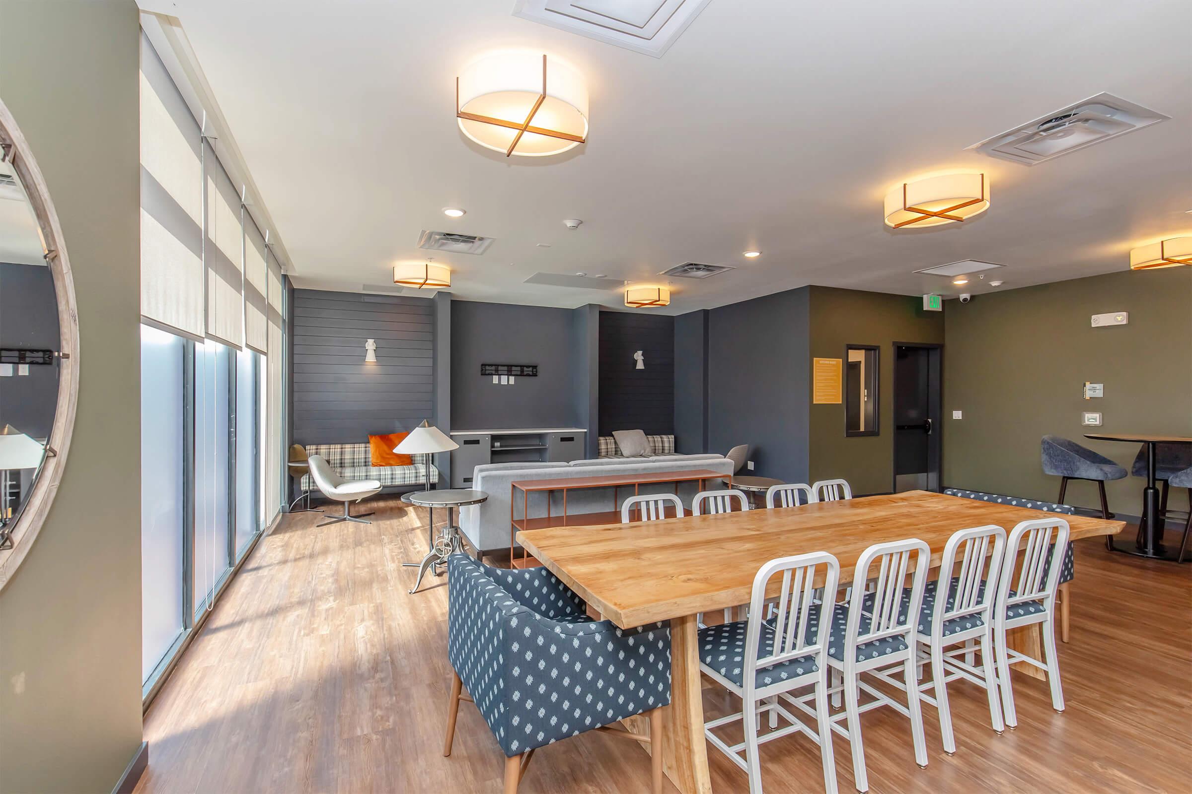 A modern, spacious lounge area featuring a large wooden dining table with white chairs, a cozy sitting area with a gray couch and armchairs, and large windows that let in natural light. Stylish lighting fixtures hang from the ceiling, contributing to a contemporary and inviting atmosphere.