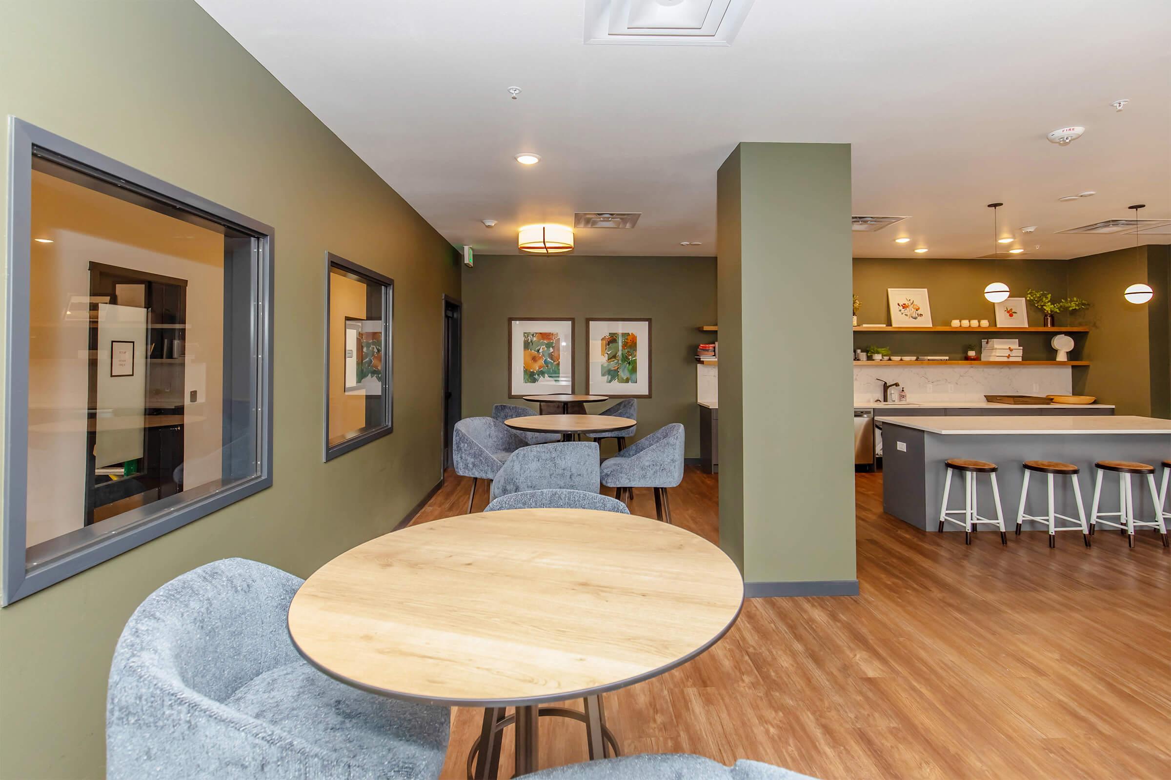 A modern lounge area with two wooden round tables surrounded by soft gray chairs. The walls feature a calming olive green color, and a kitchen area with barstools and decorative plants can be seen in the background. Framed artwork adds an inviting touch to the space.