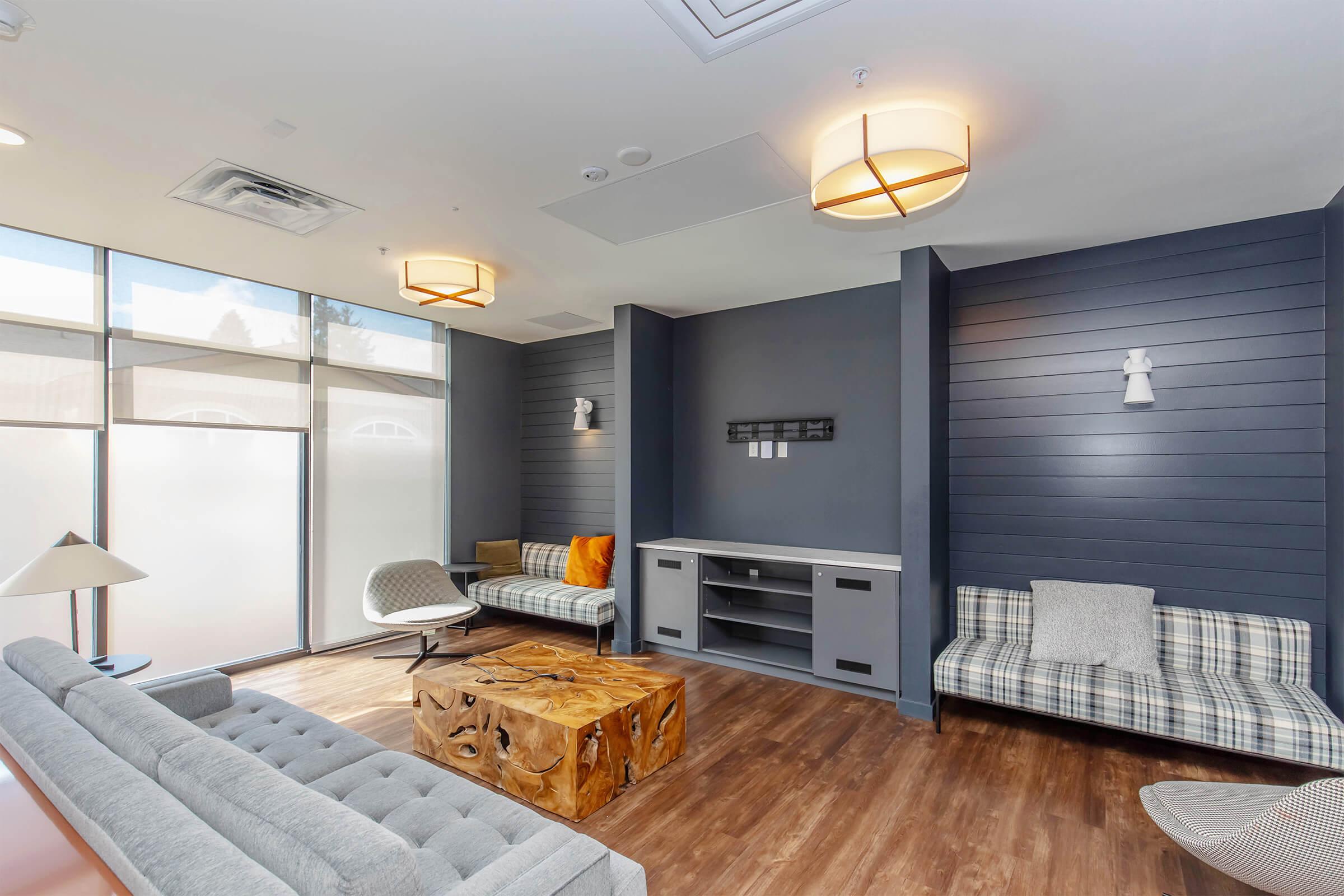 A modern lounge with two stylish sofas, a wooden coffee table, and a TV console. Soft lighting from pendant lamps and wall sconces illuminates the room. Large windows allow natural light to enter, enhancing the comfortable and inviting atmosphere.