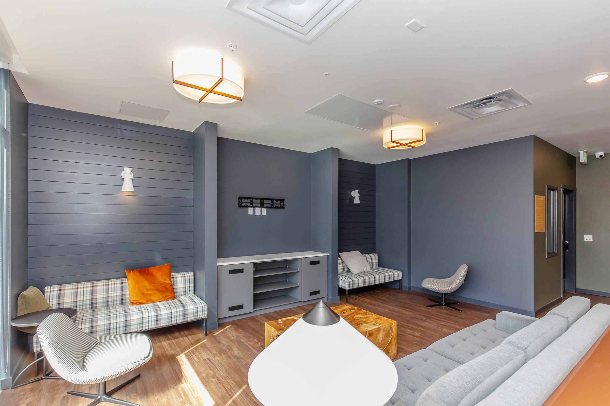 A modern lounge area featuring stylish furniture, including a gray sofa, patterned seating, and contemporary light fixtures. The walls are painted in shades of blue and gray, with warm wood accents on the floor, creating an inviting and comfortable atmosphere.