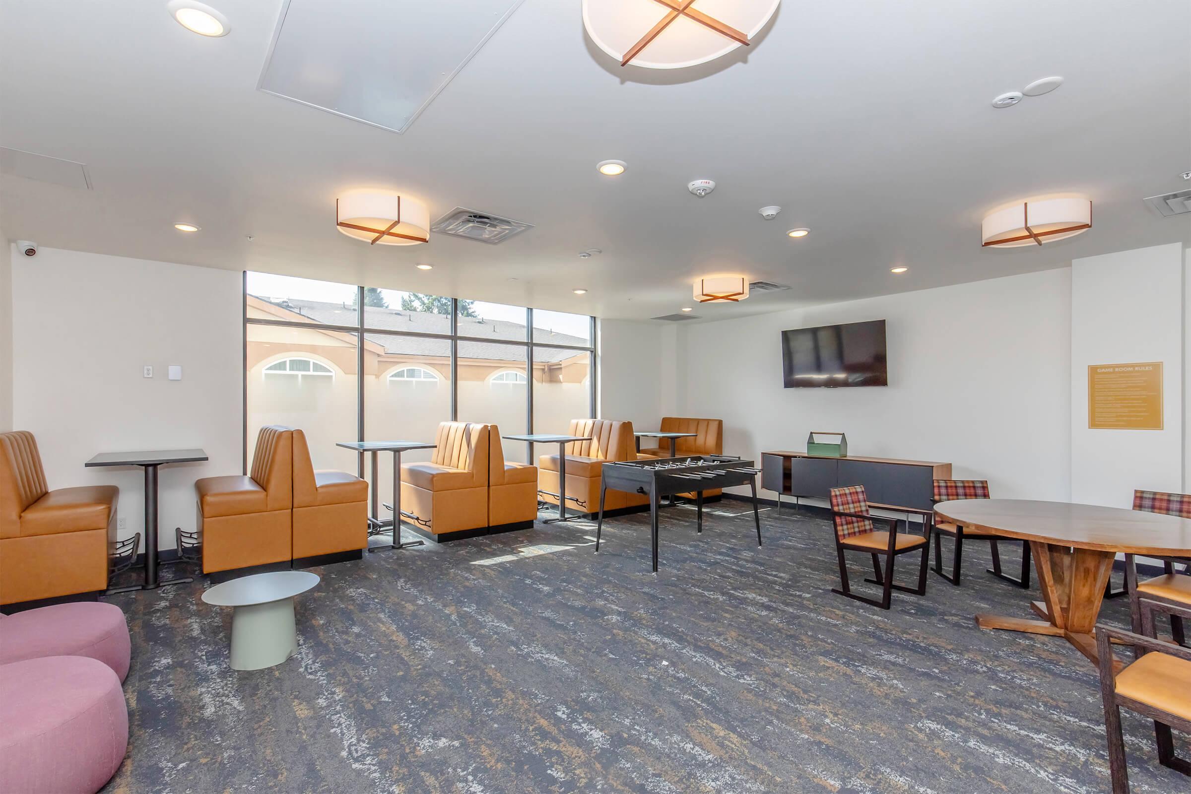 A modern lounge area featuring a mix of seating options, including brown leather chairs and light purple sofas. Large windows provide natural light, and there's a flat-screen TV mounted on the wall. Small tables are scattered around, with some multi-purpose seating near the windows.