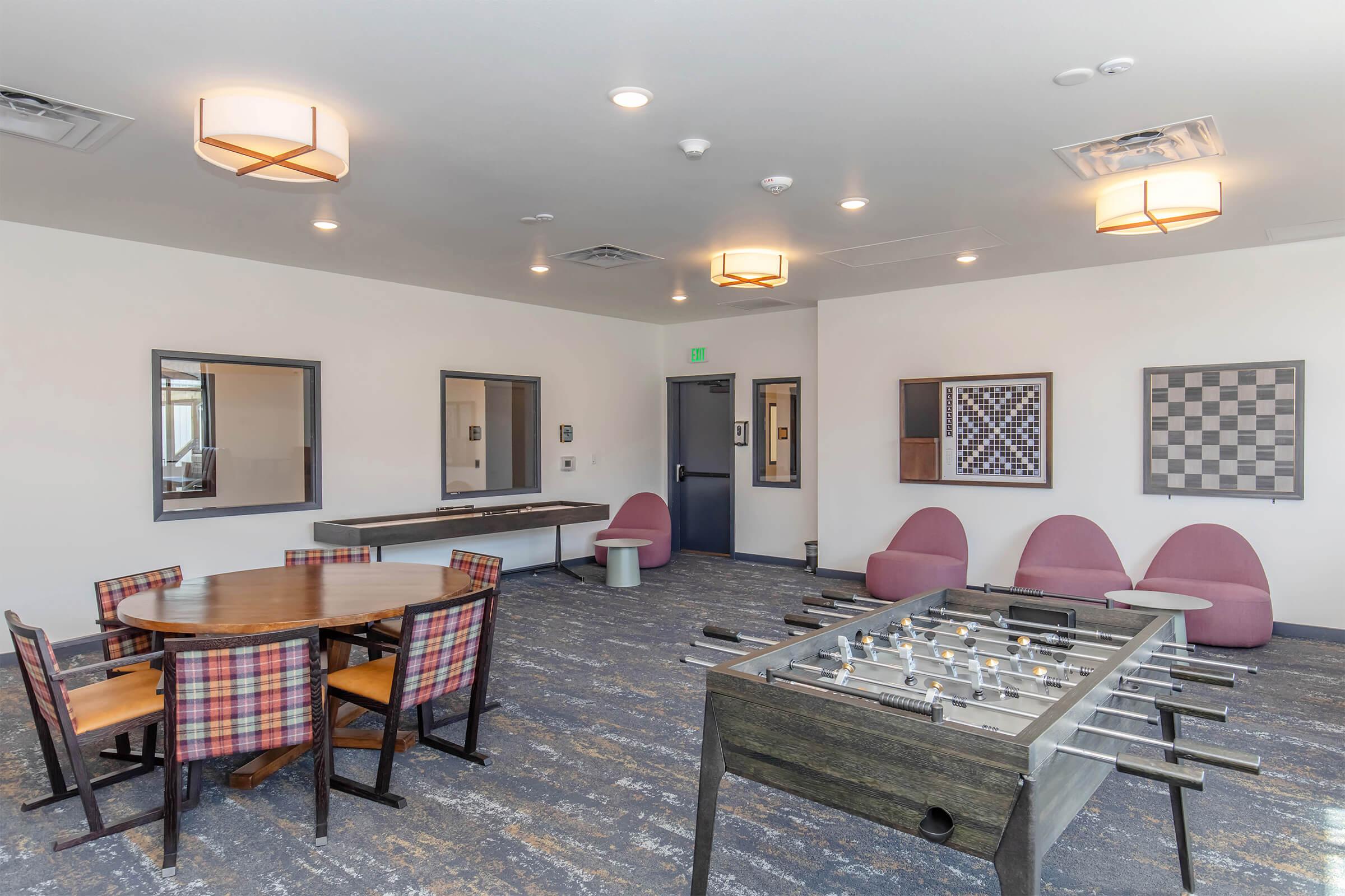 A modern recreational room featuring a large round table with plaid chairs, a foosball table, and pink upholstered lounge chairs. Two framed artworks and mirrors adorn the walls, along with a checkerboard game on display. The space is well-lit with contemporary light fixtures and has a cozy atmosphere.