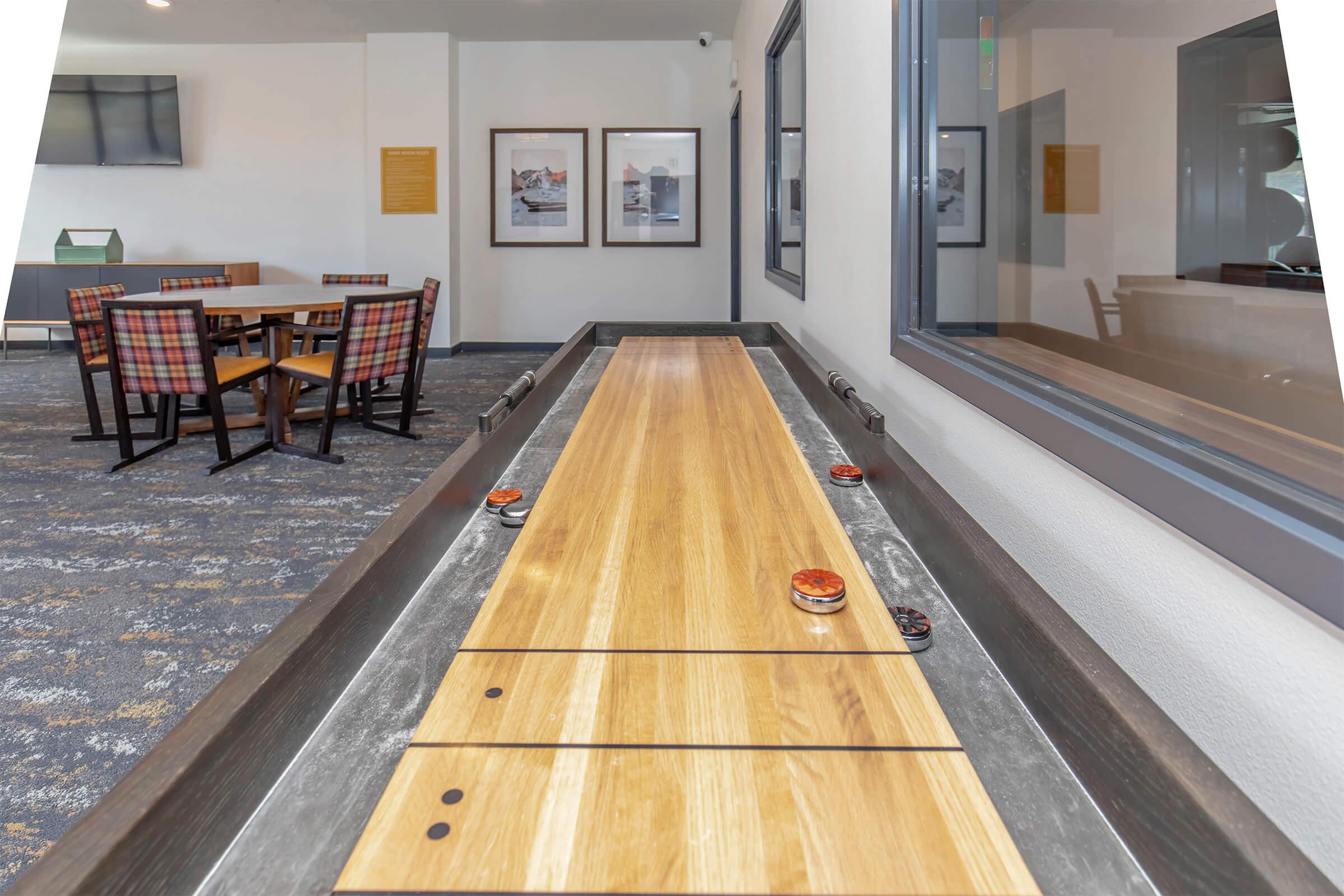 A shuffleboard table with smooth wooden surface and pucks, set in a bright room featuring tables and chairs in the background. There are framed pictures on the walls and large windows allowing natural light to fill the space.