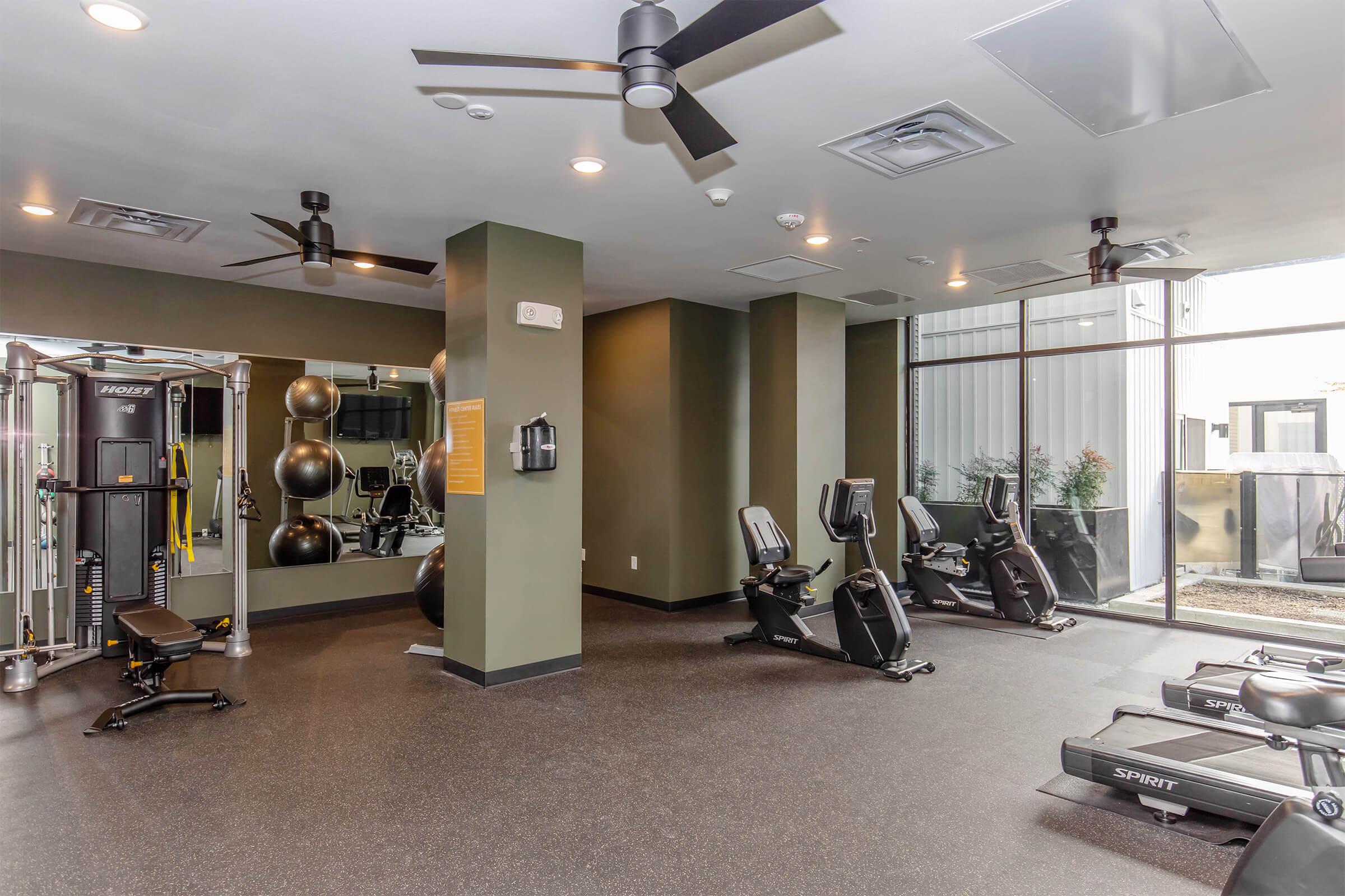A modern gym interior featuring exercise equipment, including stationary bikes, a weight machine, and fitness balls. The space is well-lit with large windows and ceiling fans, creating an inviting atmosphere for workouts. The floors are dark and the walls are painted in neutral tones.