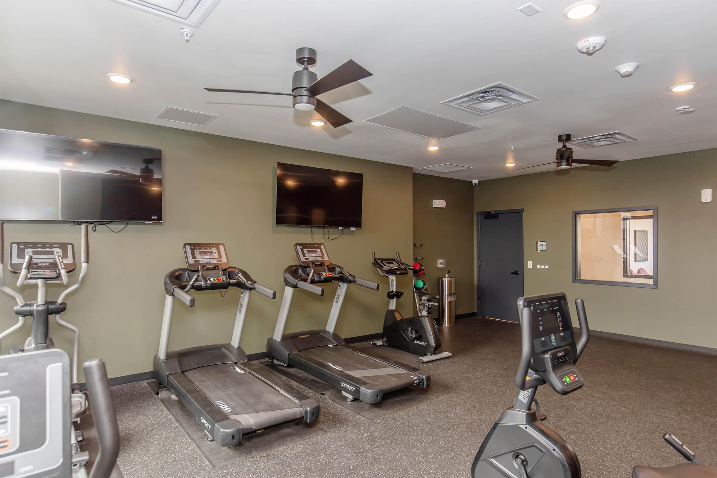 A modern gym interior featuring three treadmills and one stationary bike. The walls are painted in a subtle green color, and there are two wall-mounted televisions. The flooring is dark and the ceiling has overhead lighting and fans, creating a well-lit workout environment.