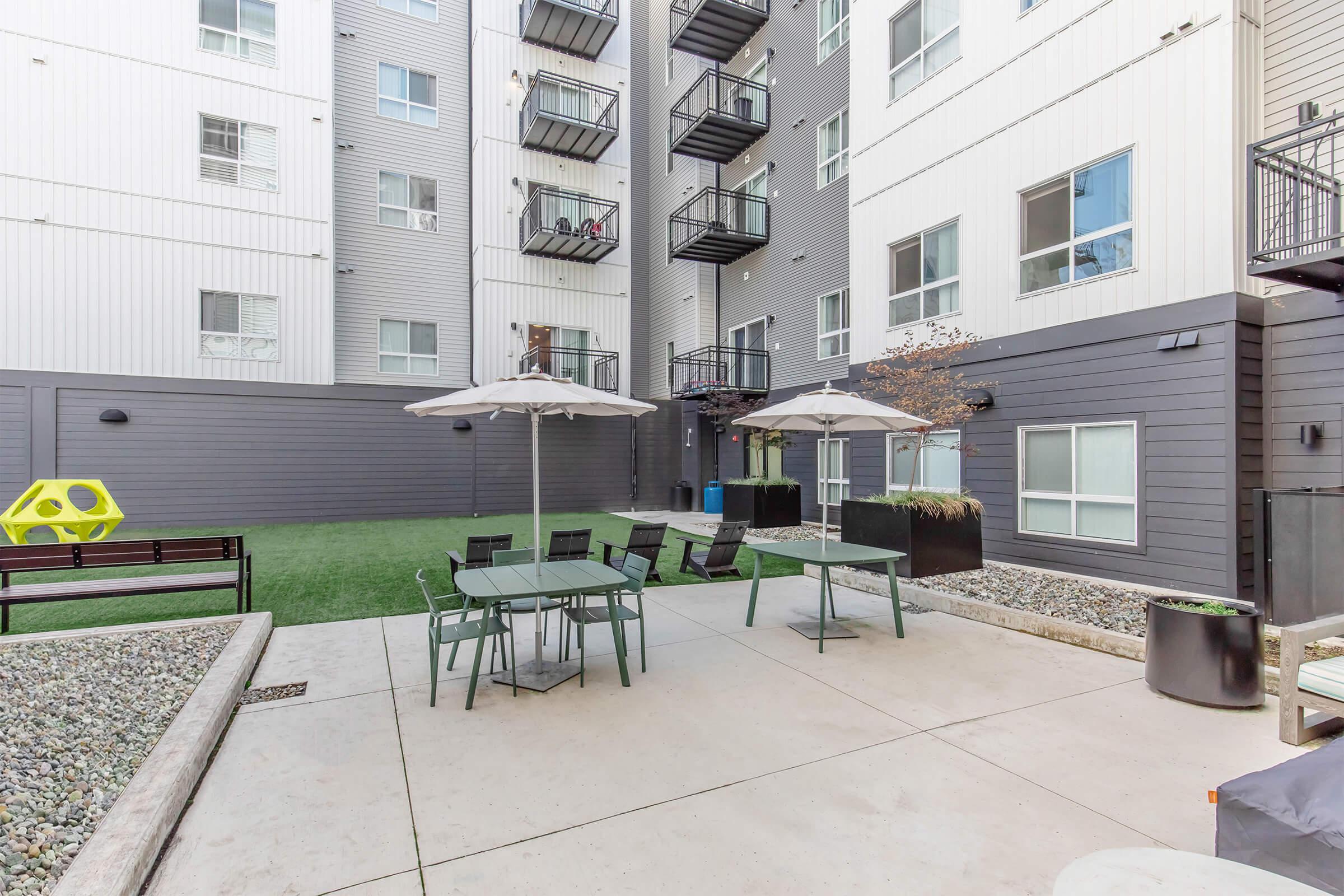 Outdoor patio area with a green lawn, featuring a modern dining set with a table and chairs under two umbrellas, surrounded by gravel and plants. In the background, there are multi-story apartment buildings with balconies.
