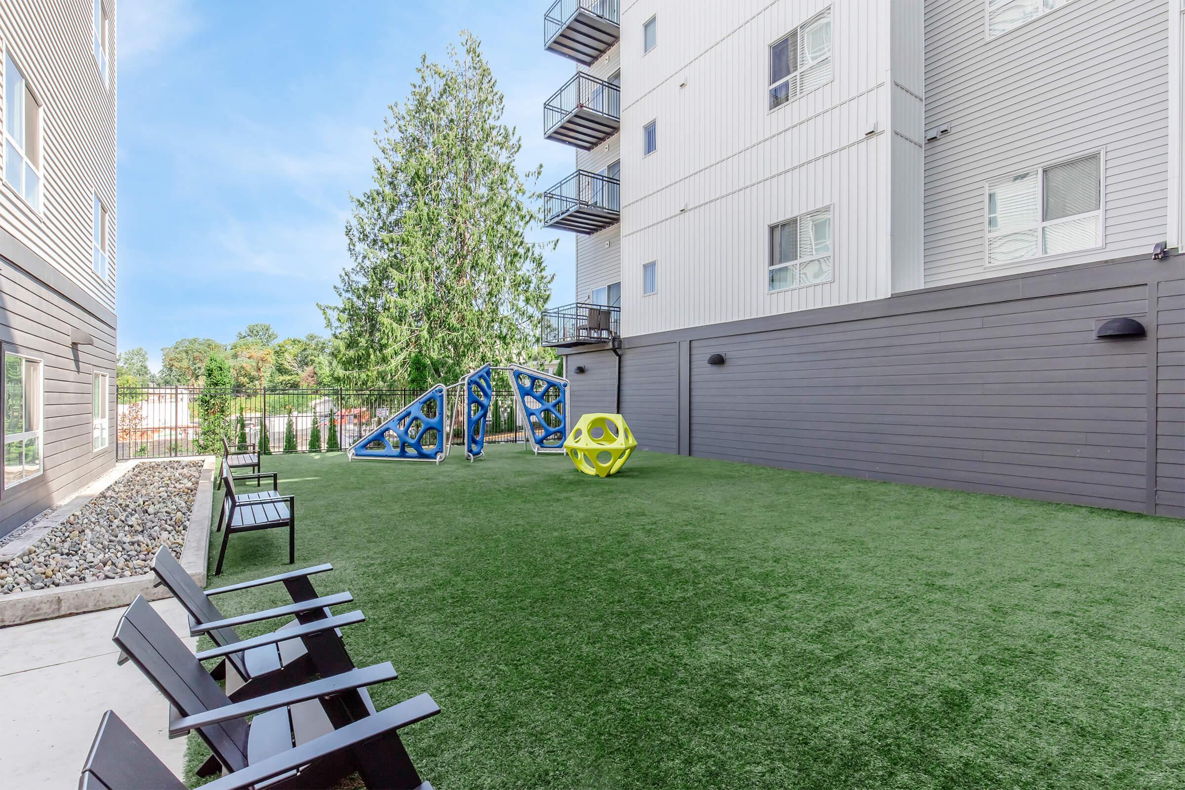 A spacious outdoor area featuring artificial grass, colorful play structures, and modern seating. The background includes a multi-story building with balconies and a tree, creating a pleasant recreational space for residents and children.