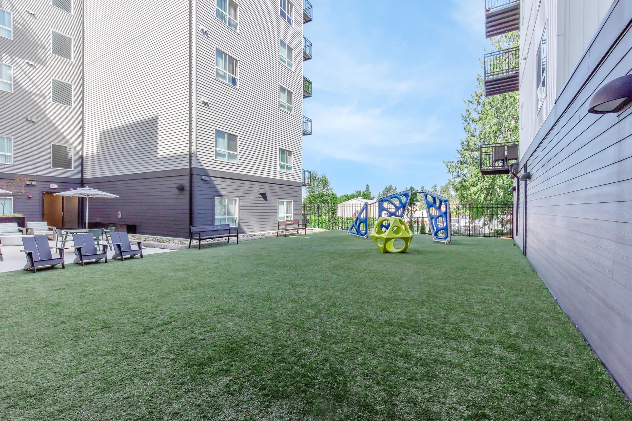 A landscaped outdoor area featuring artificial turf, with a recreational play structure in bright colors. In the background, modern apartment buildings can be seen, along with seating areas and umbrellas for relaxation. The scene is well-lit and inviting, perfect for outdoor activities.