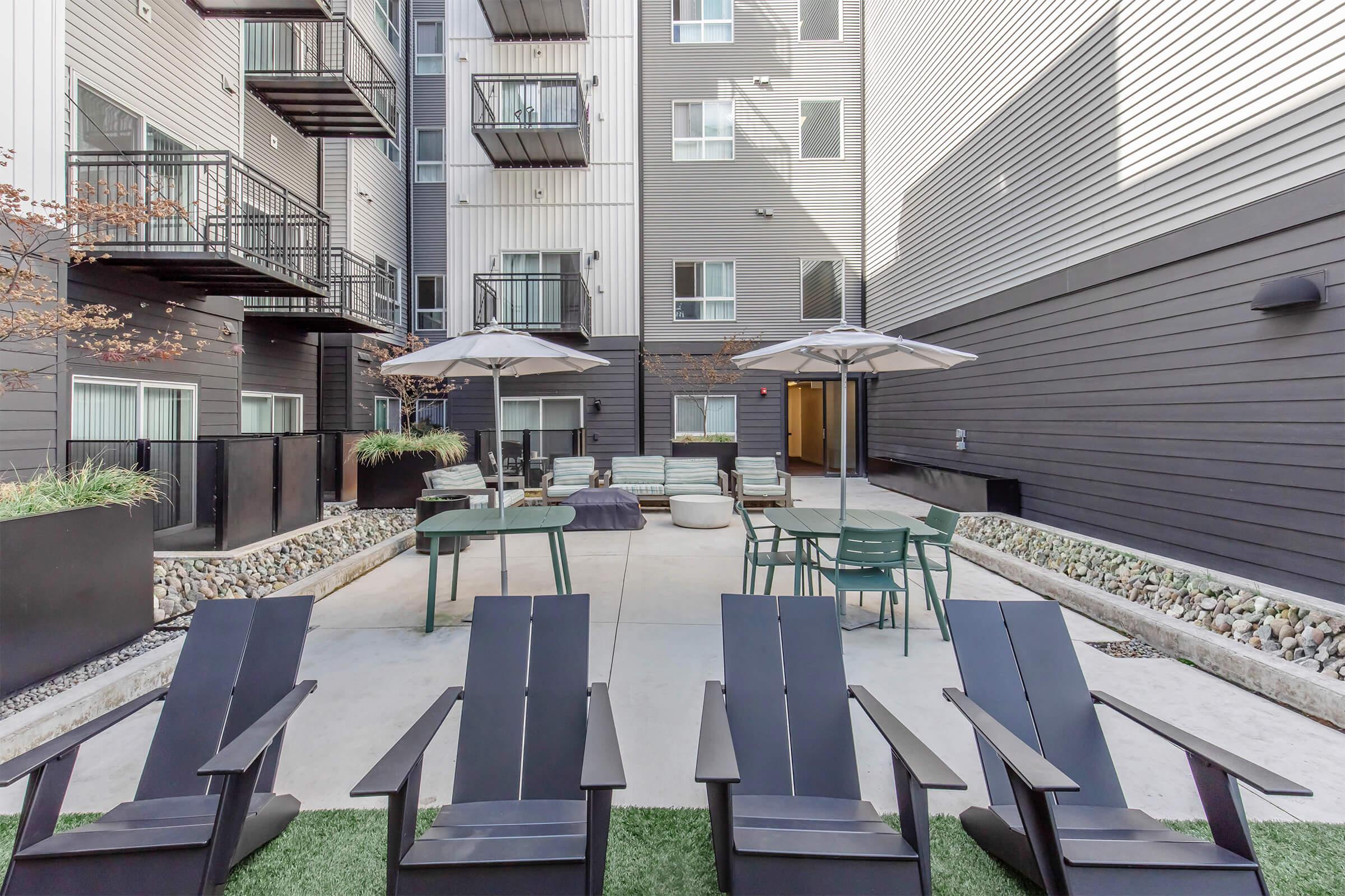 Patio area of an apartment complex featuring several black Adirondack chairs, round tables with umbrellas, and modern outdoor furniture. The ground is paved with concrete and bordered by decorative stones. Surrounding buildings have multiple windows and modern architecture. Trees provide hints of greenery.