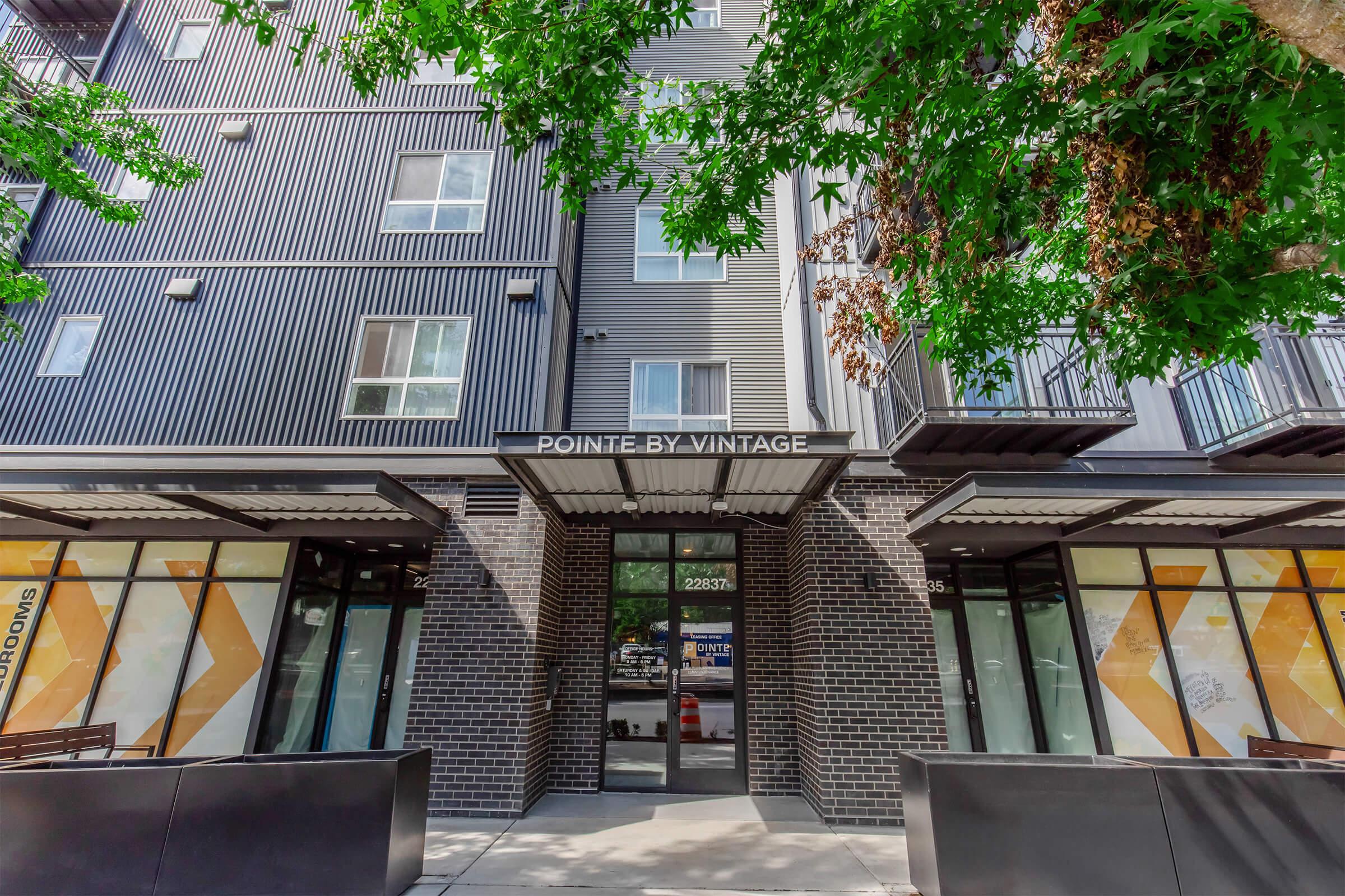 Modern apartment building with a sleek design, featuring dark paneling and large front windows. The entrance displays "POINTE BY VINTAGE" overhead, surrounded by greenery and brick accents. There are balconies on the upper floors, and street-level seating areas visible on either side.