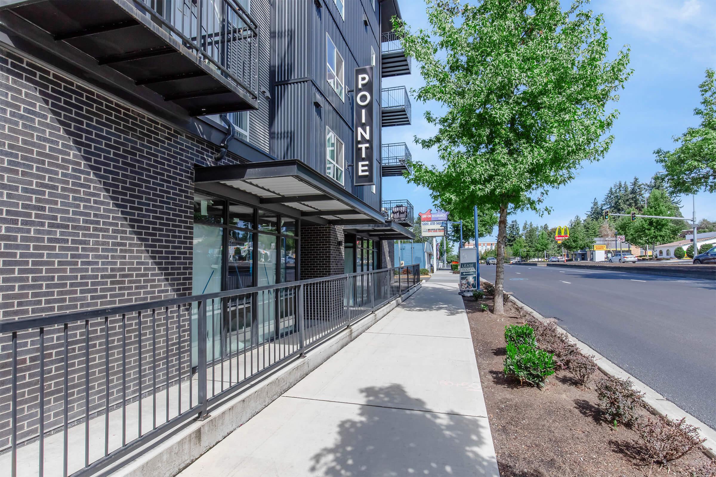 A sidewalk next to a modern building featuring black brick, large windows, and the name "POINTE" displayed prominently. There are trees along the sidewalk and a road visible in the background with a few cars. The scene is bright and well-lit, suggesting a pleasant day.