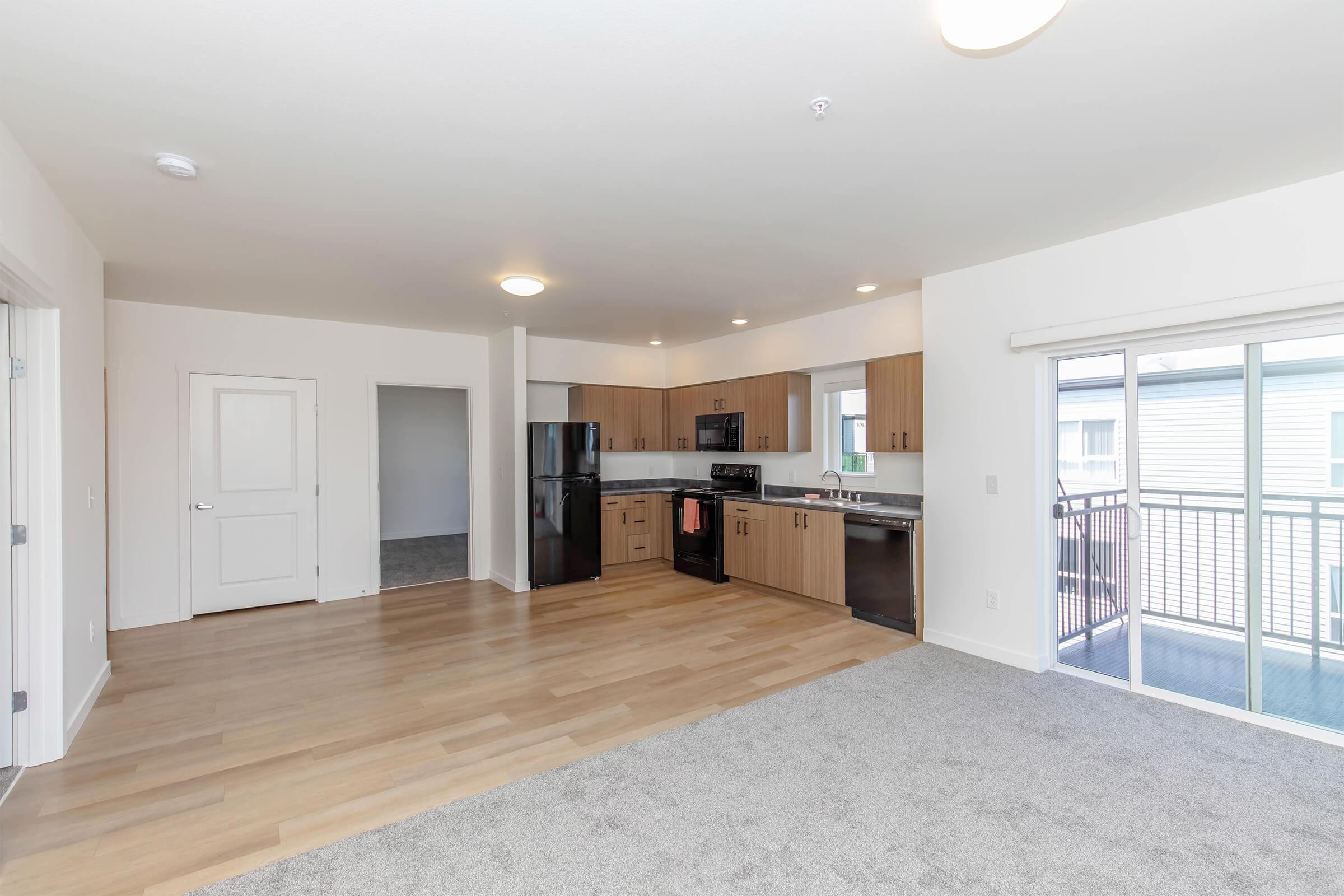 A spacious living area featuring light wood flooring and grey carpet. The modern kitchen is equipped with dark cabinets, black appliances, and a contrasting light countertop. A sliding glass door leads to a small balcony, and there is a doorway to another room in the background. Natural light fills the space.