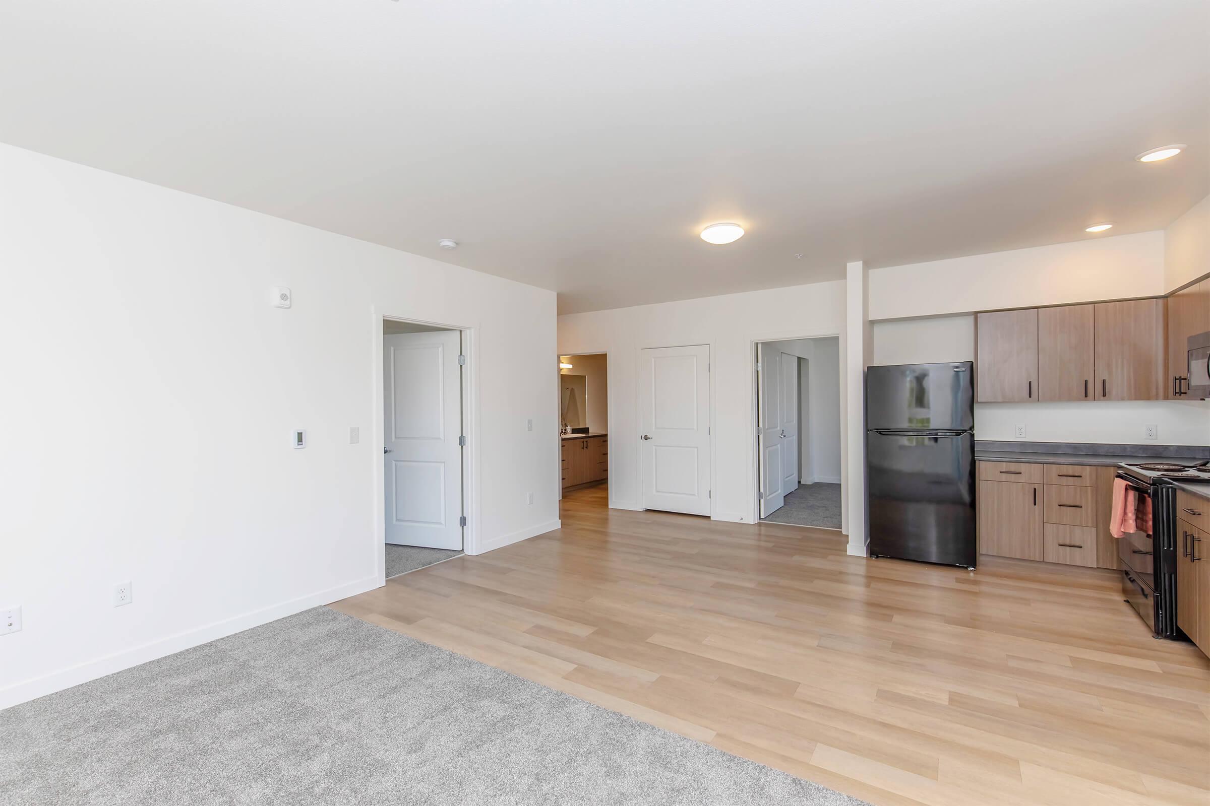 A spacious, modern apartment interior featuring an open layout. The room has light-colored walls, wooden flooring, and a large carpeted area. An adjacent kitchen is visible with wooden cabinetry and a black refrigerator. Two doors lead to other areas, enhancing the airy ambiance.