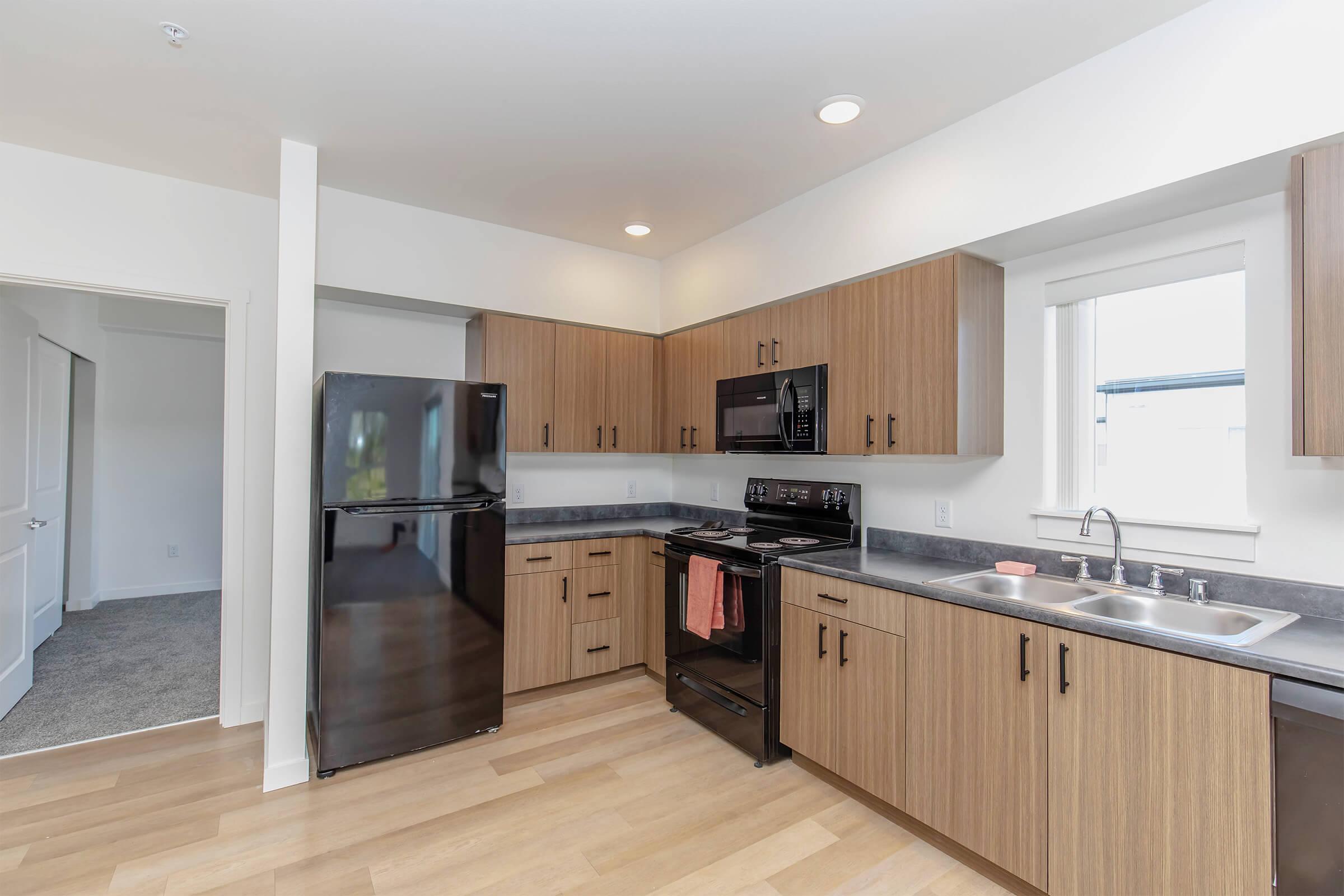 A modern kitchen featuring wood cabinets, stainless steel appliances, a black refrigerator, and a stove. Natural light comes through the window above the sink, and the flooring is light wood. An adjacent room is visible through an open door, suggesting a connected living space.